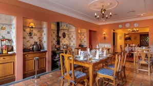 Uma área de refeições com mesas e cadeiras de madeira, um candelabro e uma parede de azulejos com motivos de frutas.