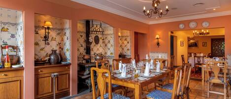 A dining area with wooden tables and chairs, a chandelier, and a tiled wall with fruit patterns.