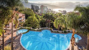 A large, circular swimming pool surrounded by palm trees and lounge chairs, with a view of a cityscape and mountains in the background.