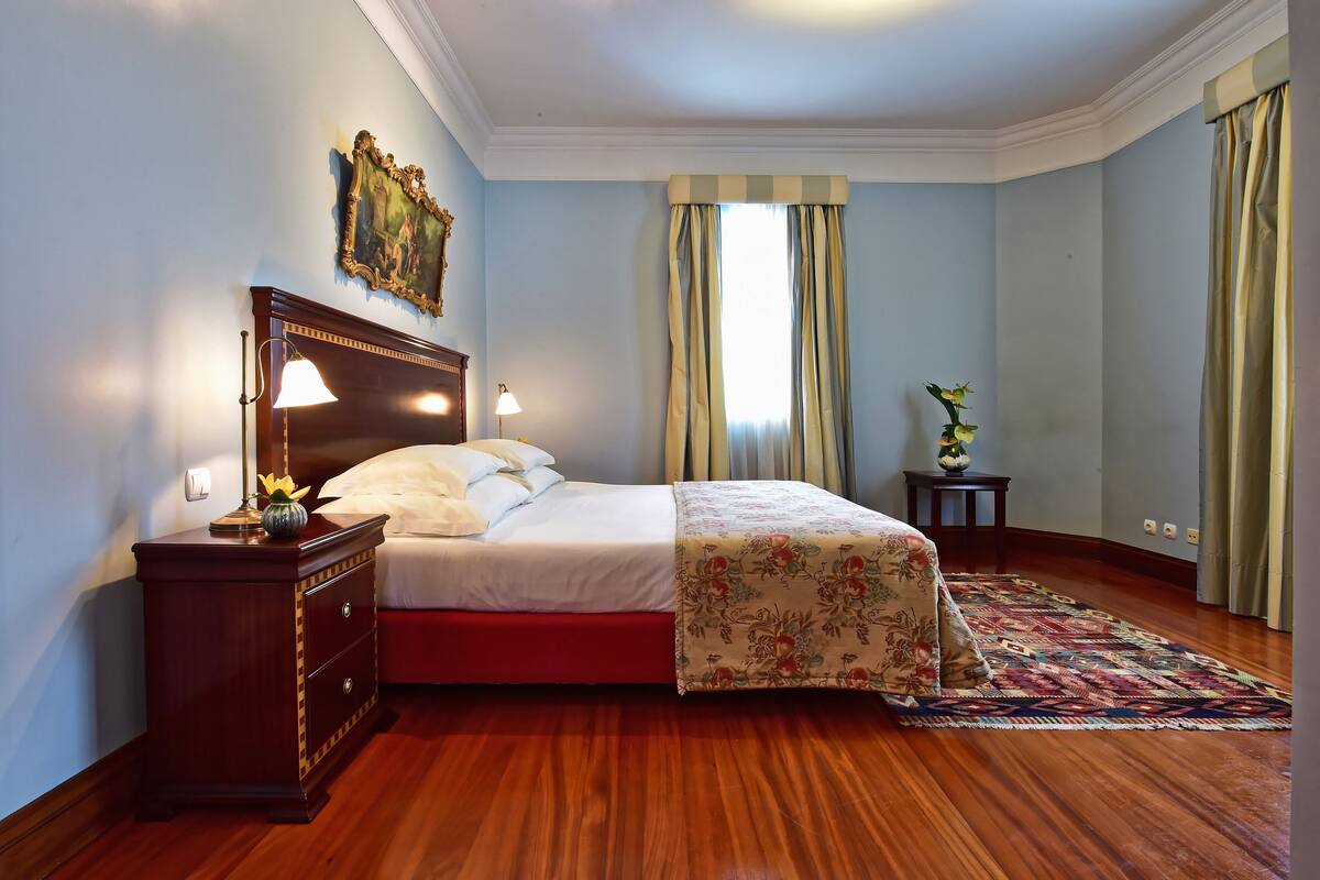 Une chambre avec un plancher en bois, un lit recouvert d’une couvre-lit à motifs floraux, une table de chevet avec une lampe, un tableau au mur et une fenêtre avec des rideaux.