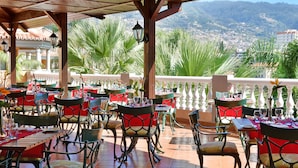 Restaurante com área externa, cadeiras e mesas vermelhas, com vista para a cidade e colinas.