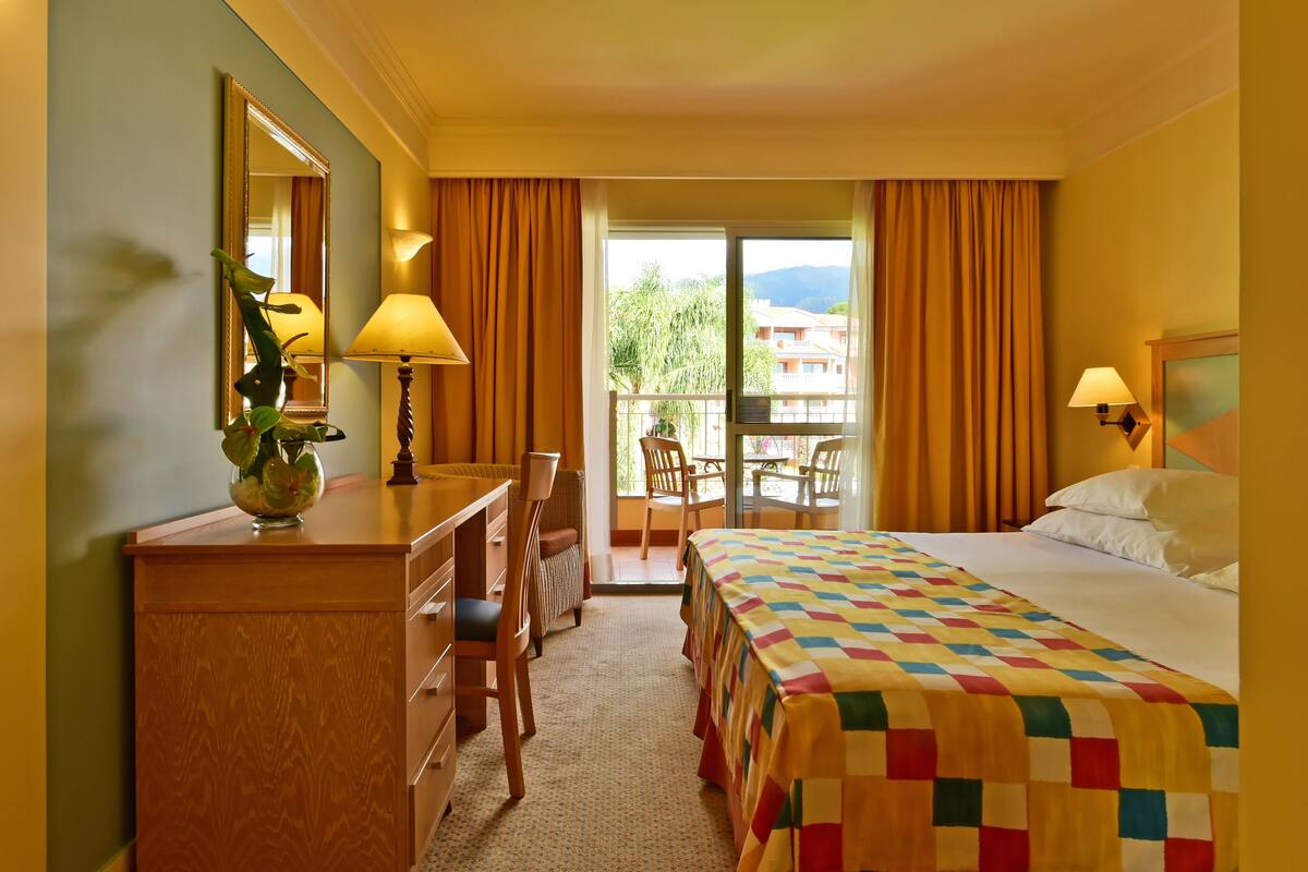 Une chambre d’hôtel comprenant un lit, un bureau avec une chaise, une lampe, un miroir et une vue sur l’extérieur.