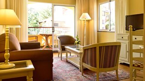 A hotel room with a sofa, armchair, dining table, lamp, TV, and a view of the outdoors. - Pestana Village Garden Hotel (Funchal)