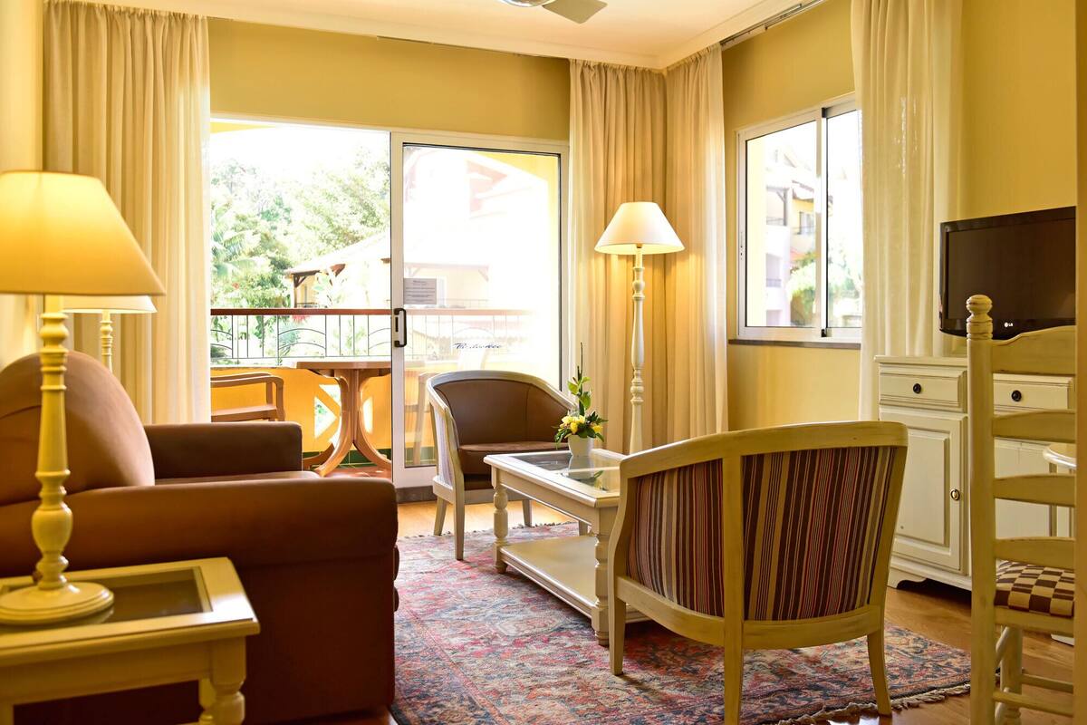 Une chambre d’hôtel équipée d’un canapé, d’un fauteuil, d’une table à manger, d’une lampe, d’une télévision et offrant une vue sur l’extérieur.