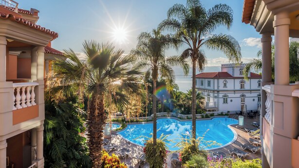 A pool area with palm trees, a sunlit sky, and a building with a red roof in the background. - Pestana Village Garden Hotel (Funchal)