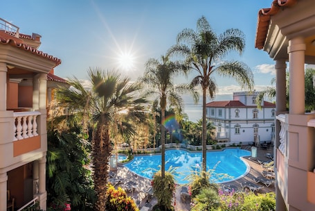 A pool area with palm trees, a sunlit sky, and a building with a red roof in the background.