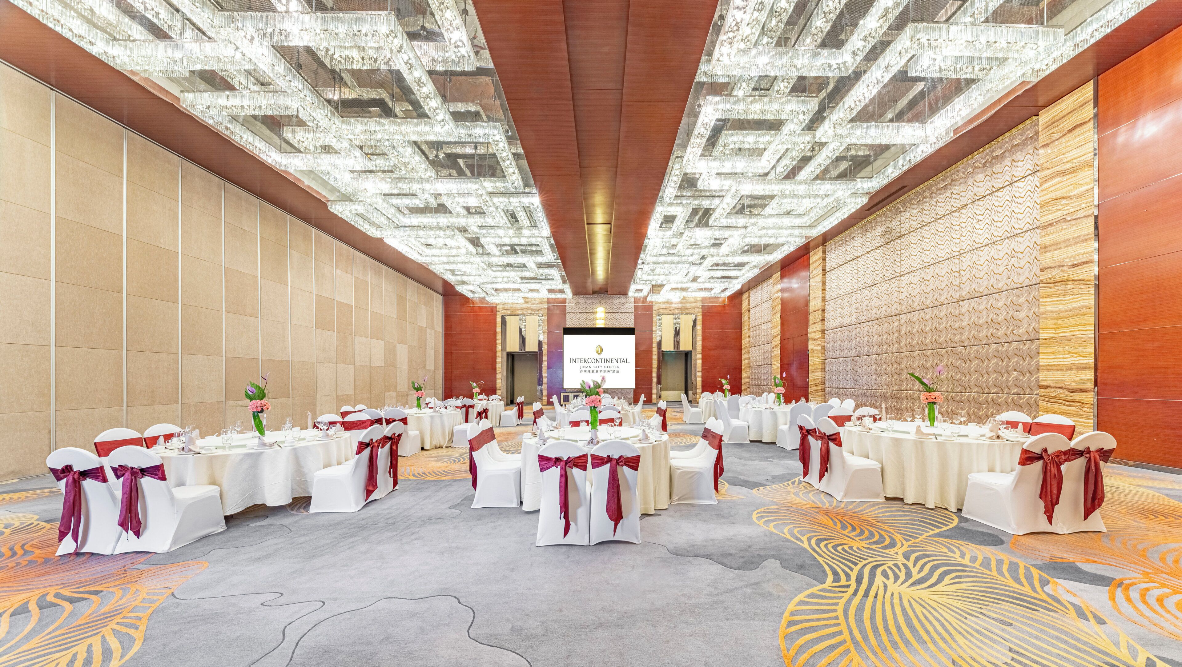 A banquet hall with round tables, white chairs, and red sashes, featuring a patterned carpet and a chandelier ceiling.