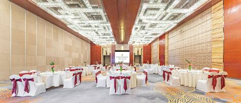 A banquet hall with round tables, white chairs, and red sashes, featuring a patterned carpet and a chandelier ceiling.