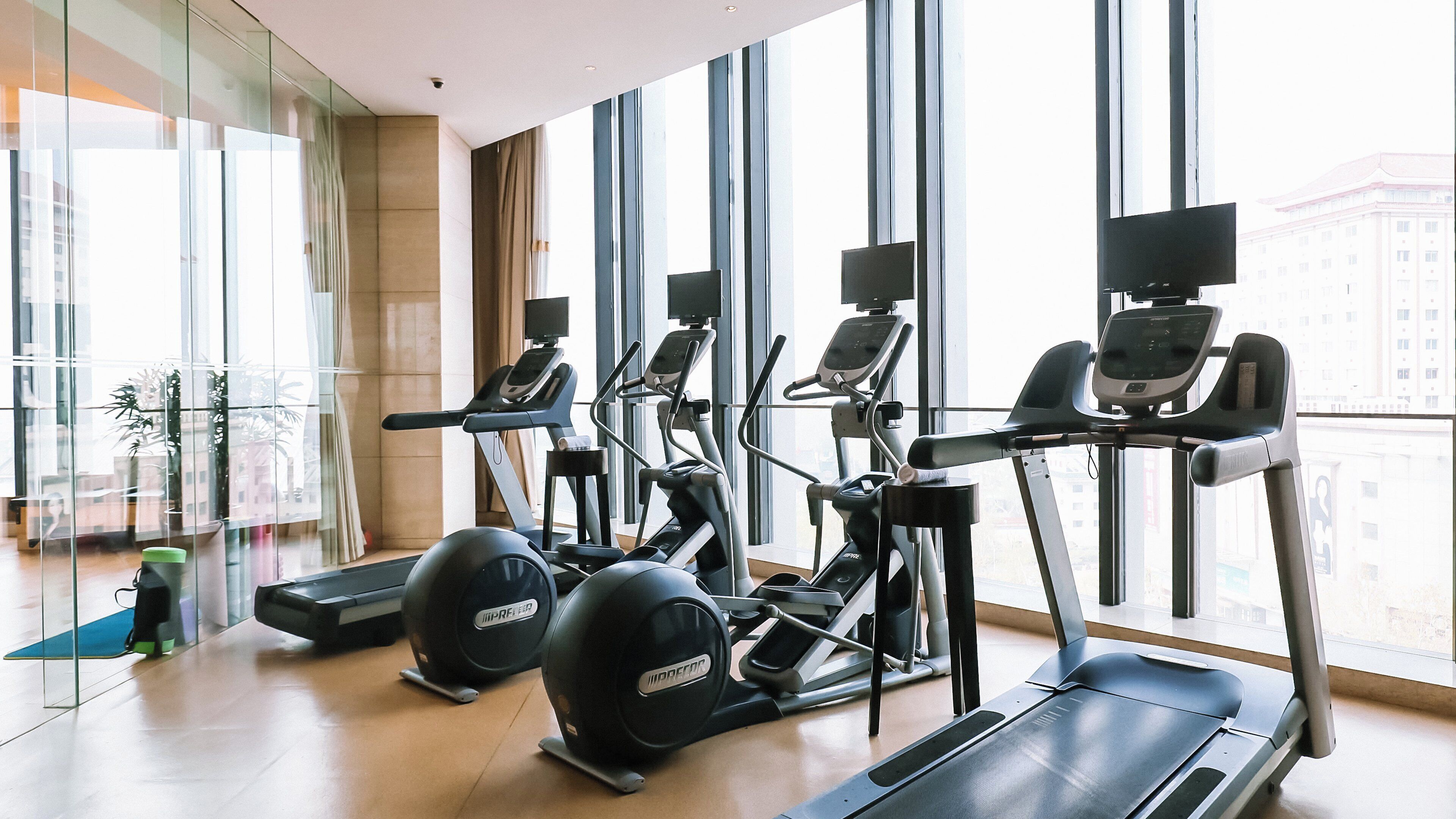 A gym with treadmills, elliptical machines, and exercise balls, large windows, and a view of buildings.