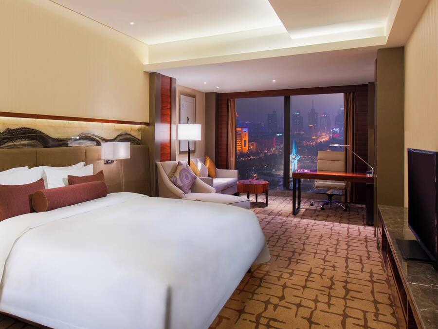 Habitación de hotel con una cama grande, un escritorio, una silla, un televisor y vistas de la ciudad de noche.