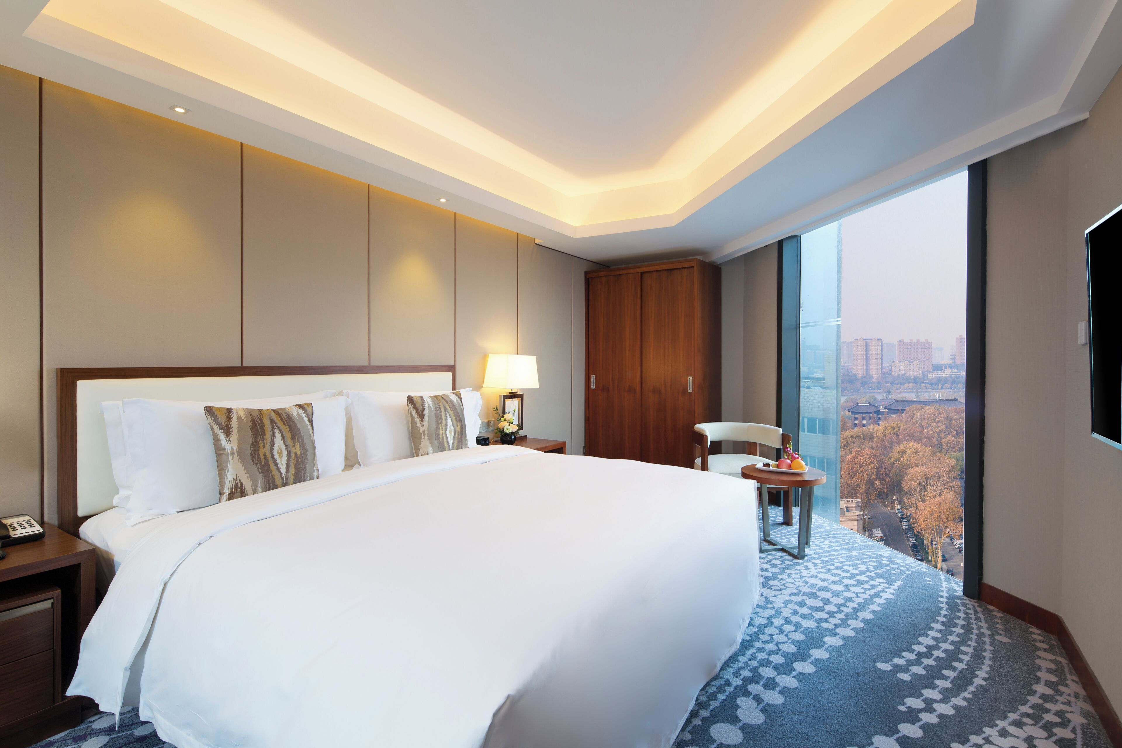 Habitación de hotel con una cama grande, un armario de madera, un escritorio con lámpara y vistas a un paisaje urbano a través de un ventanal amplio.