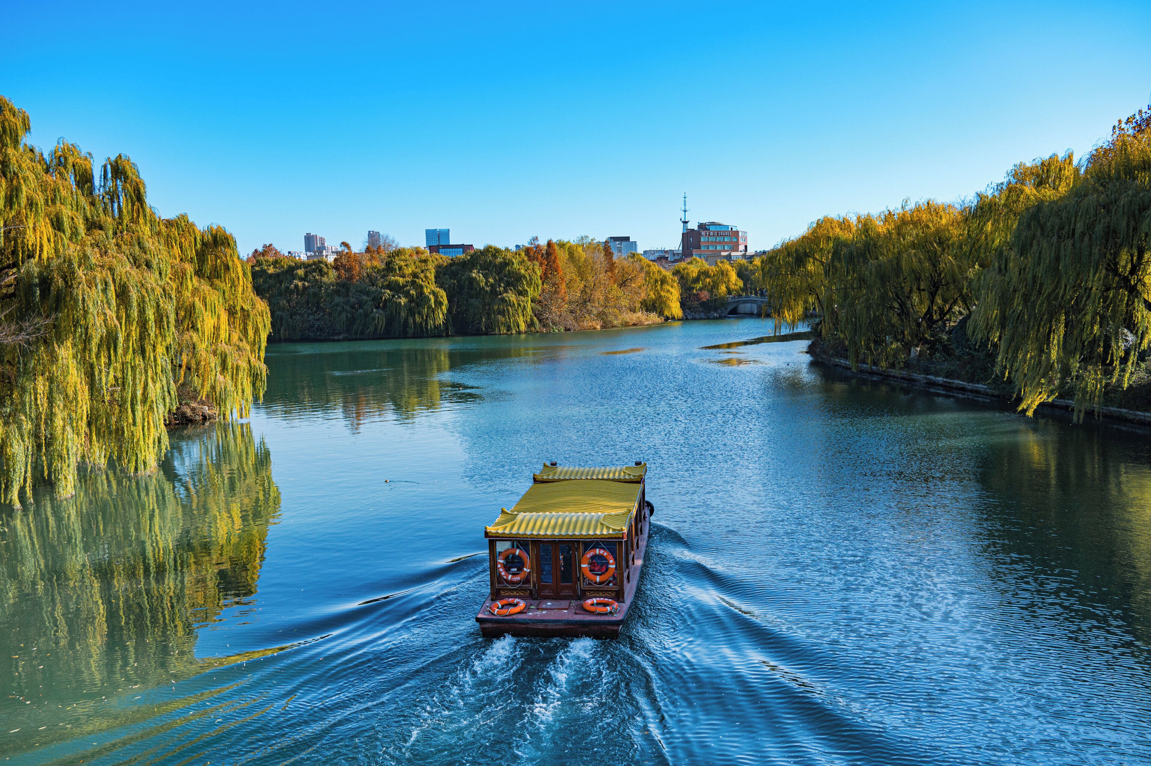 A riverboat on a calm river with trees on both sides and a city skyline in the background.