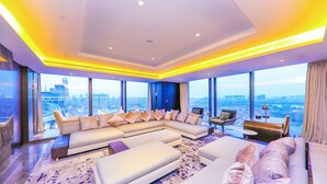 A modern hotel room with a large L-shaped sectional sofa, a small round table, and a view of the cityscape through large windows.