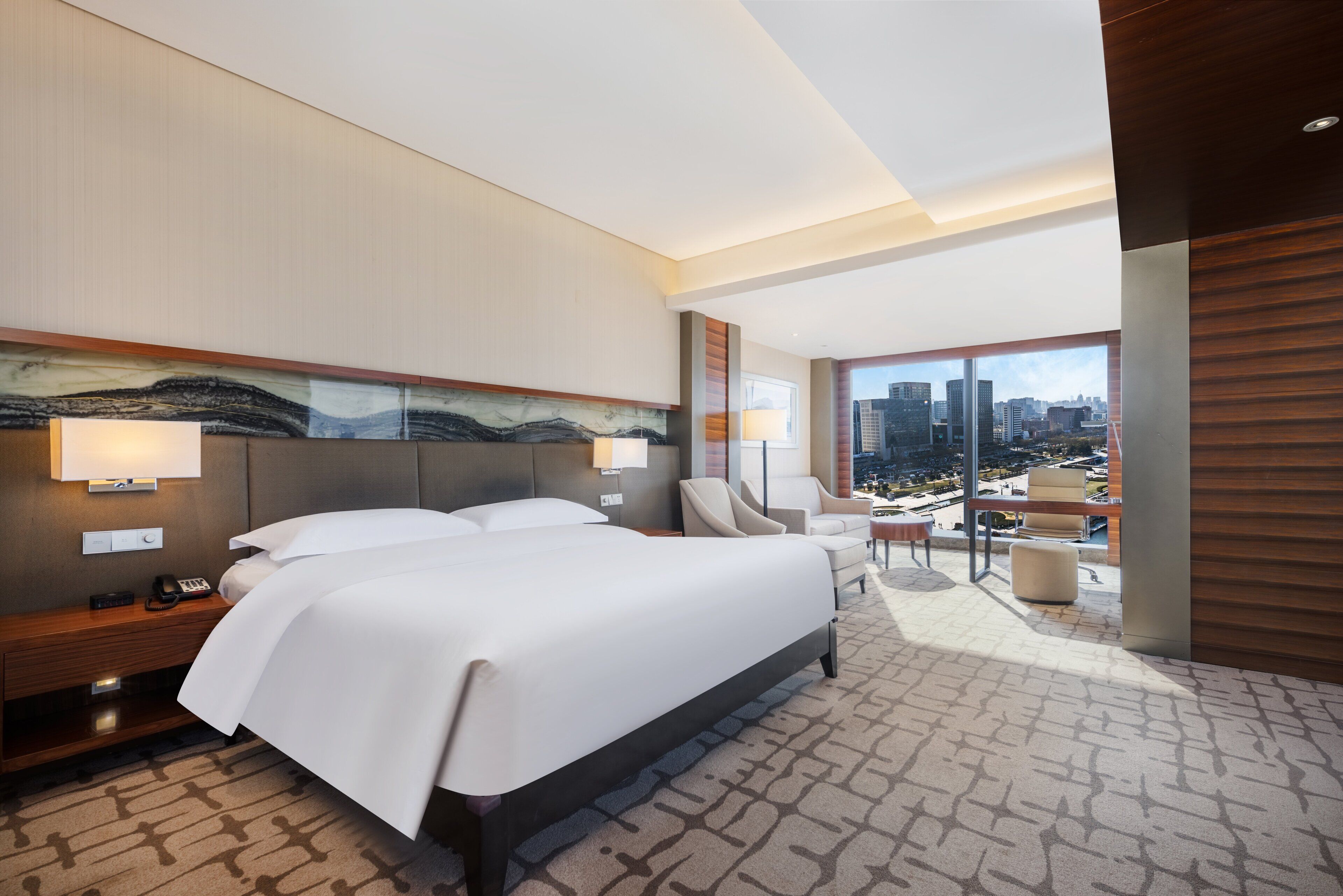 A modern hotel room with a large bed, a desk, a chair, and a view of the city through large windows.