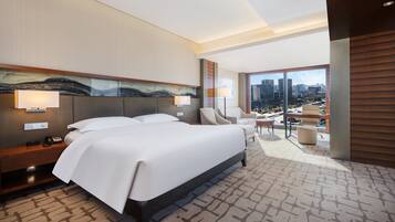 A modern hotel room with a large bed, a desk, a chair, and a view of the city through large windows.