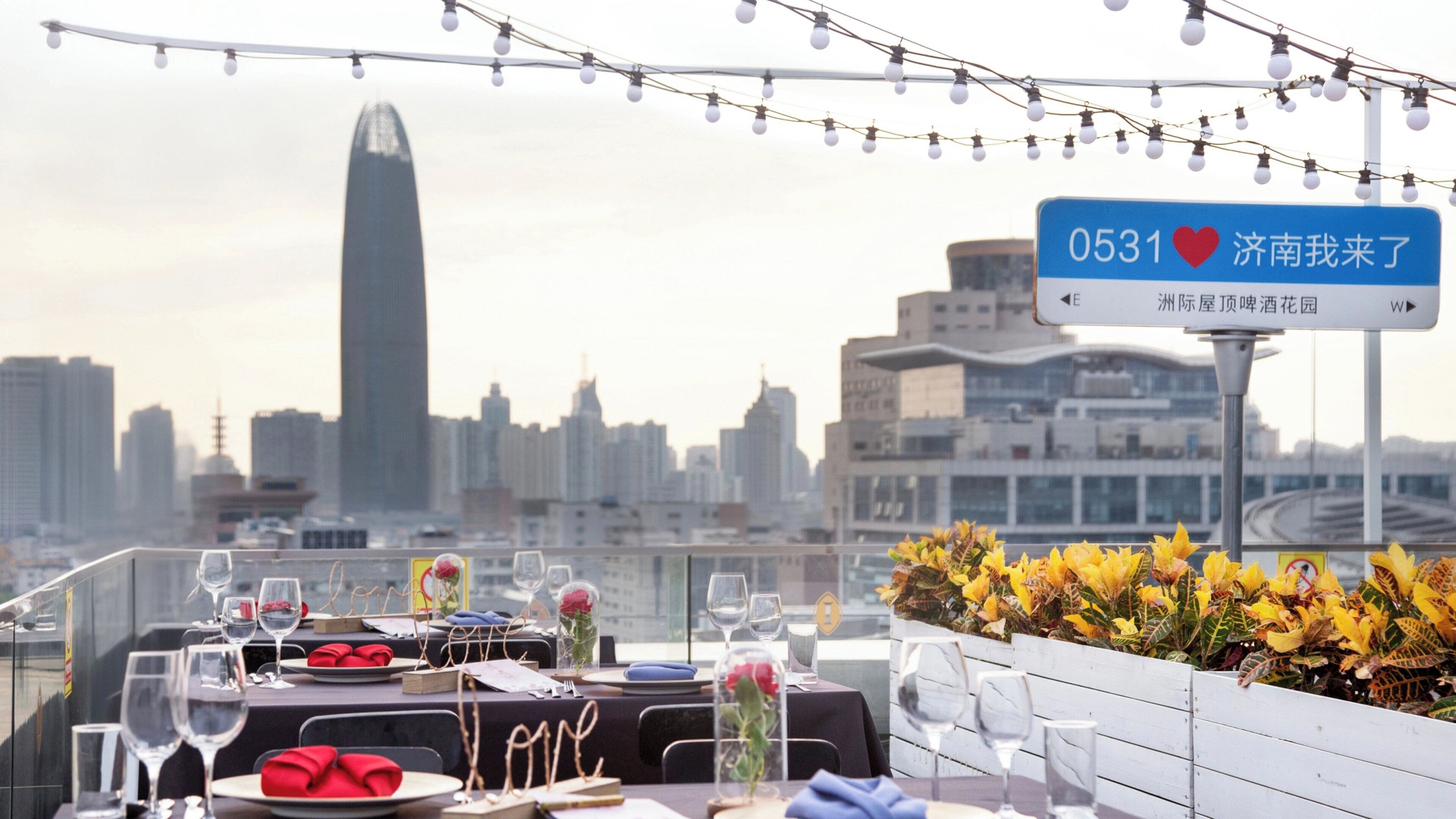A rooftop dining area with tables set for two, city skyline in the background, and a sign indicating the location.