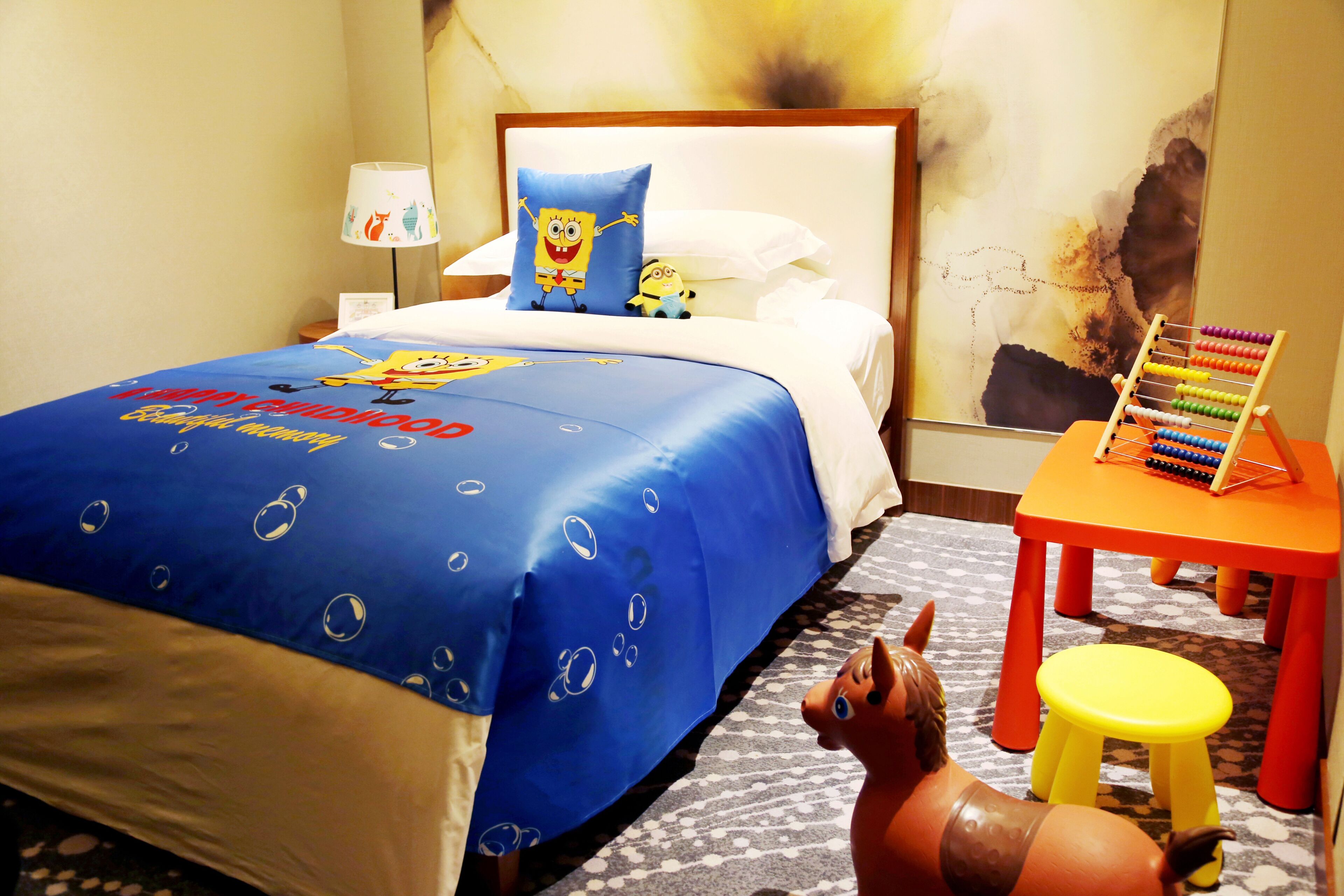 A hotel room with a bed, a nightstand, a lamp, a pillow with a cartoon character, a small table with an abacus, and a toy horse.