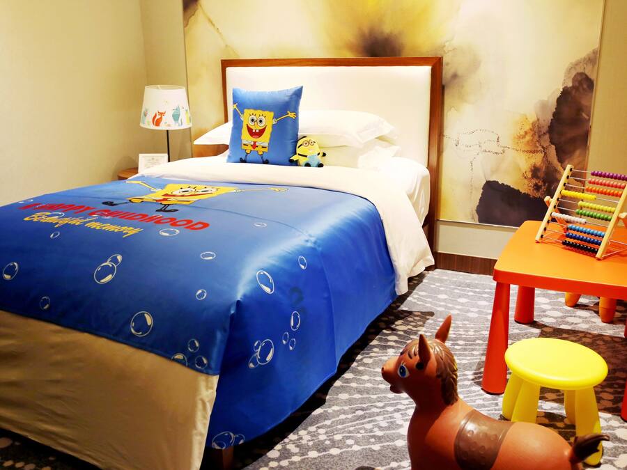 A hotel room with a bed, a nightstand, a lamp, a pillow with a cartoon character, a small table with an abacus, and a toy horse.