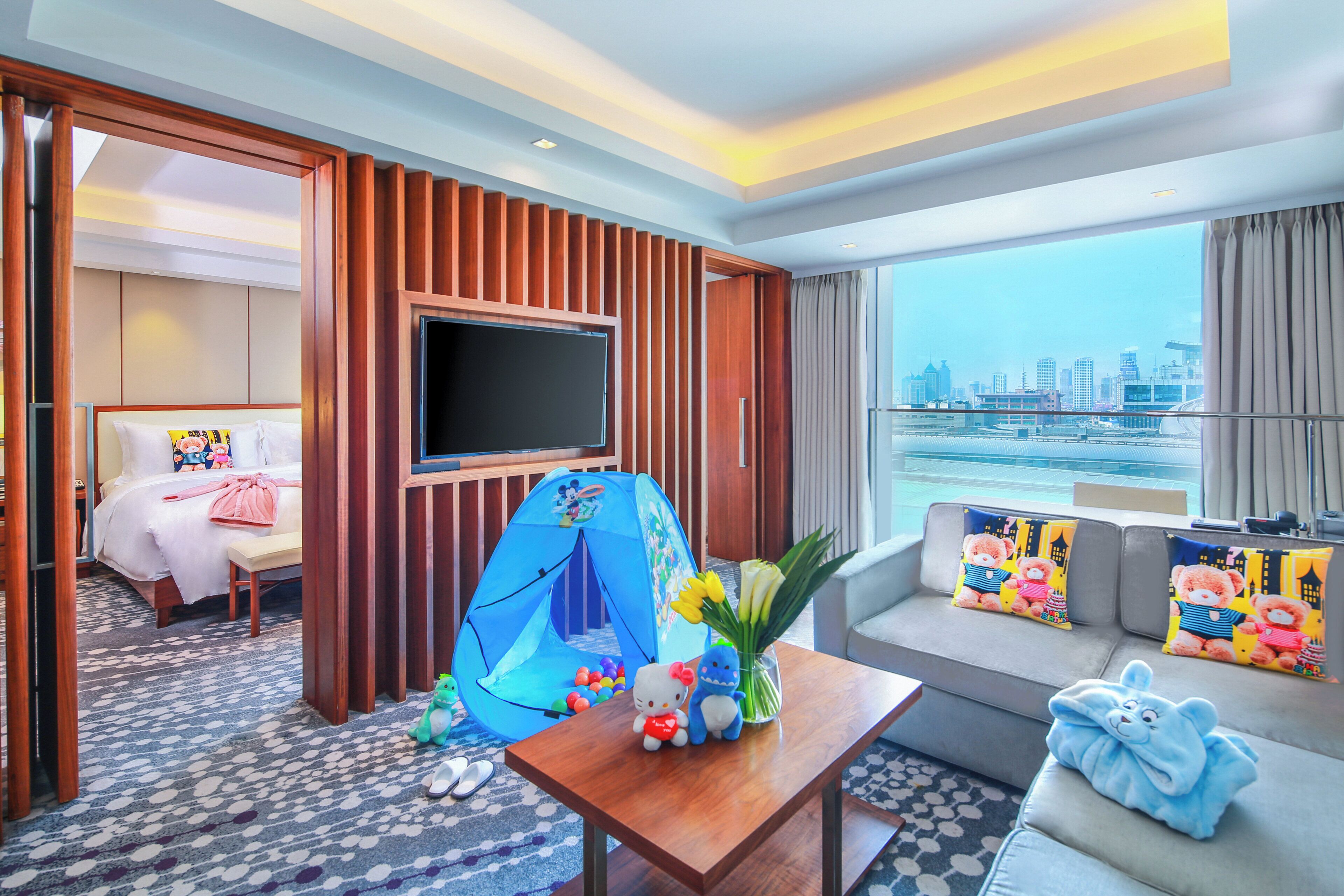 Habitación de hotel con cama, televisor, tienda de juegos, sofá con cojines con motivos de osos y vista a la ciudad.