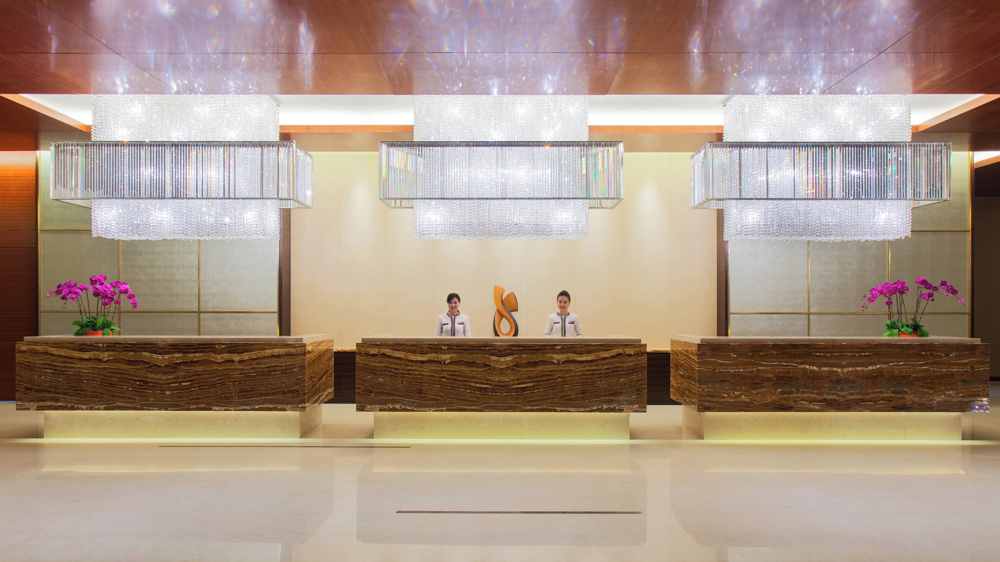 A reception desk with three counters, each with a marble top and a potted plant. Above the counters are three large, rectangular chandeliers.
