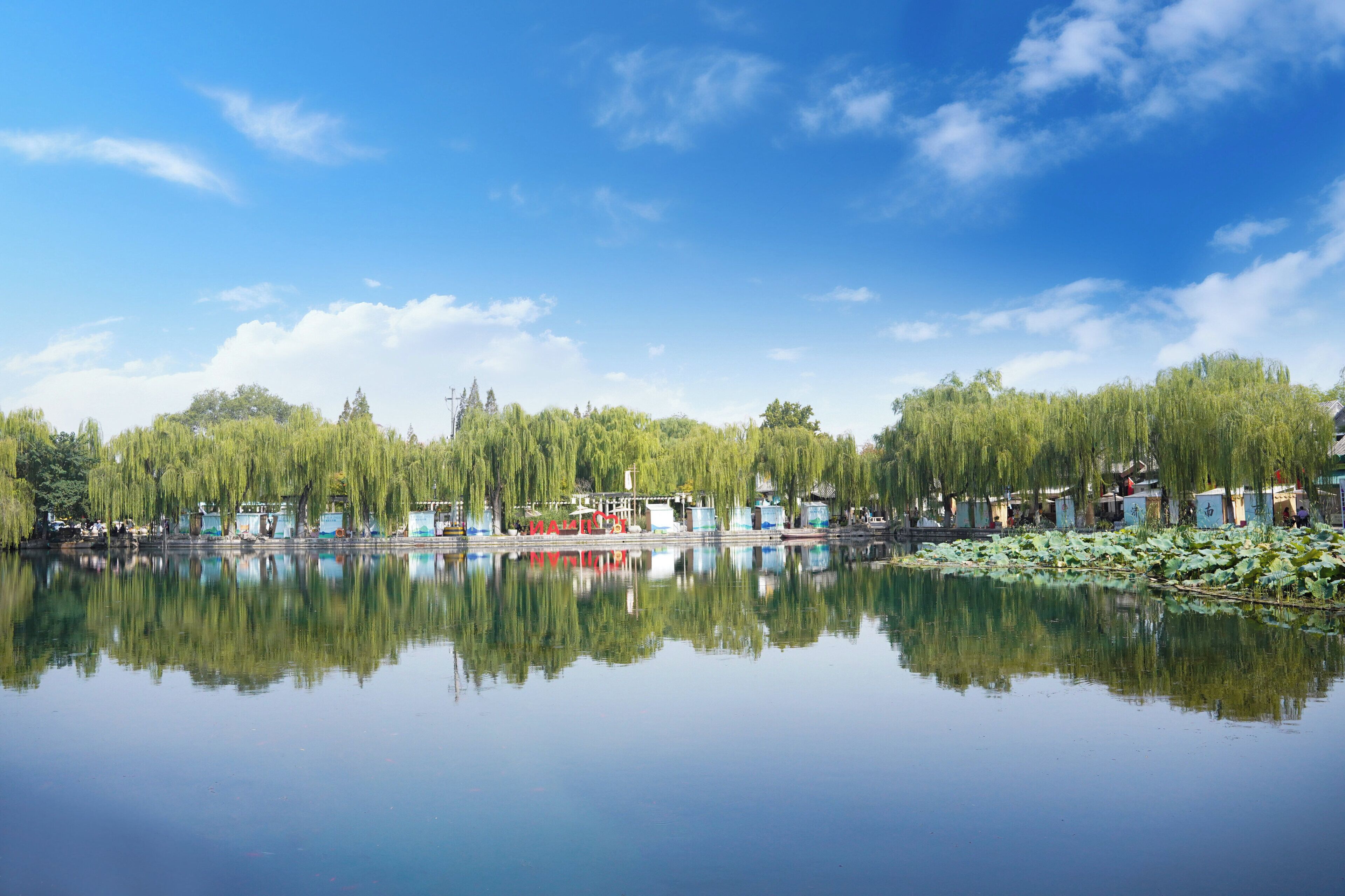 A calm lake with reflections of trees and buildings, surrounded by lush greenery and a clear blue sky.