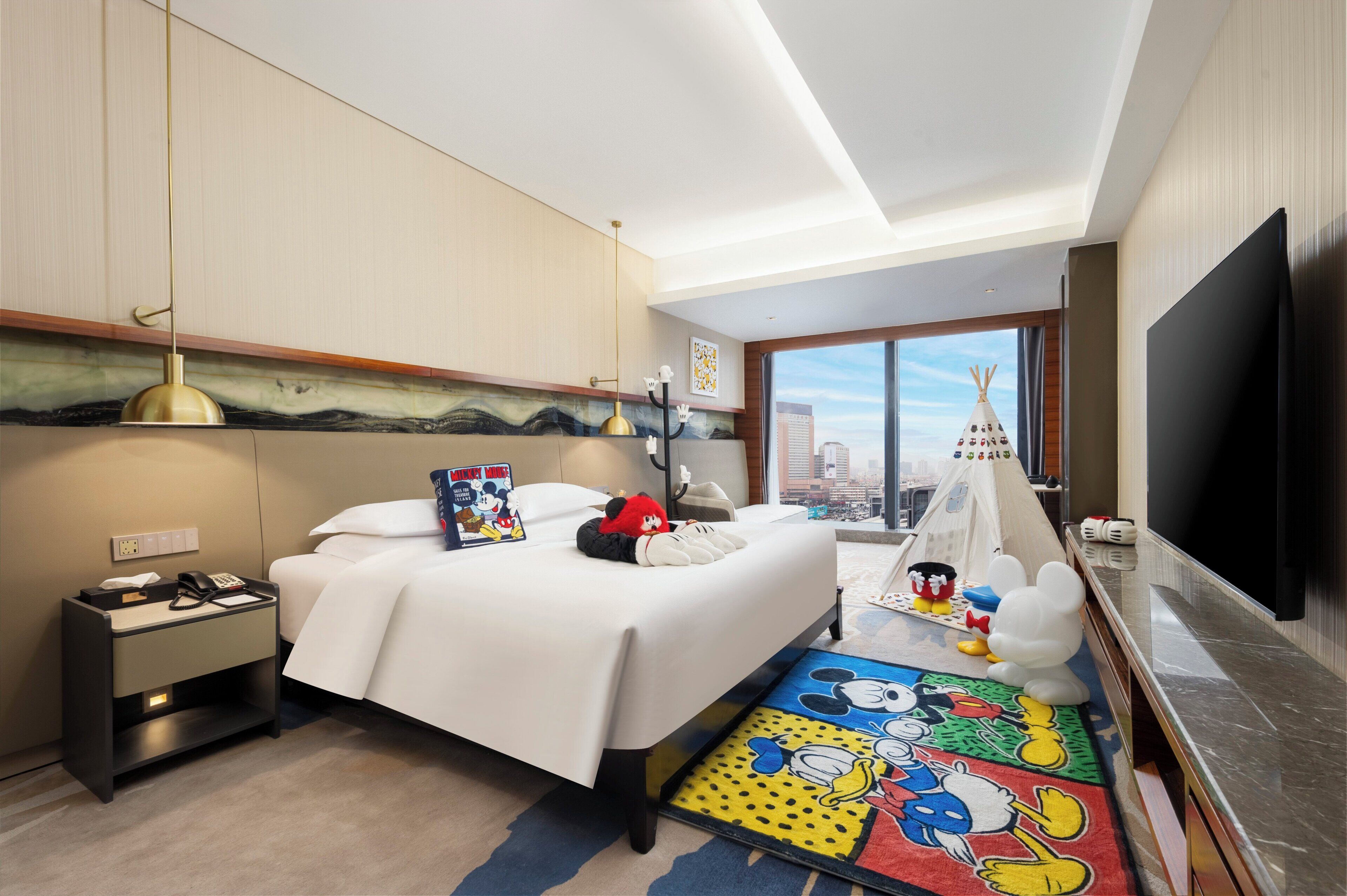 A hotel room with a bed, a nightstand, a TV, a window with a city view, and a Mickey Mouse themed rug.