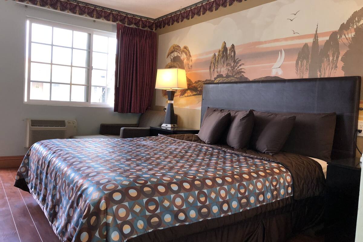 A hotel room with a large bed, a nightstand, a lamp, a window with curtains, and a mural on the wall.