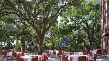 Un espace de restauration en plein air, avec des tables et des chaises, entouré d’arbres et bordé d’un bâtiment en briques.