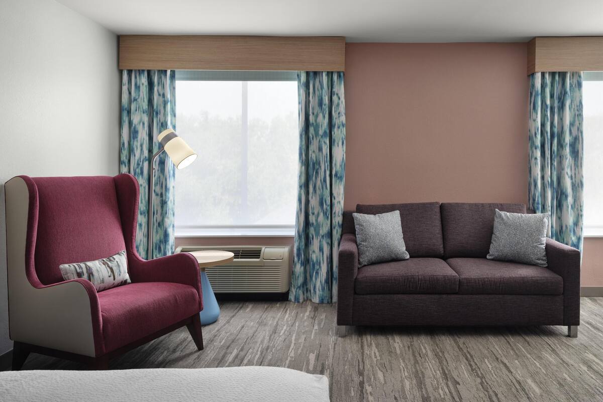 Une chambre d’hôtel avec un fauteuil rouge, un canapé marron, une lampe et une vue sur l’extérieur à travers des rideaux.