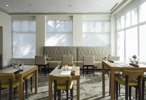 Ein Hotel-Speisesaal mit einem langen Tisch, mehreren Stühlen, einer gepolsterten Bank und großen Fenstern.