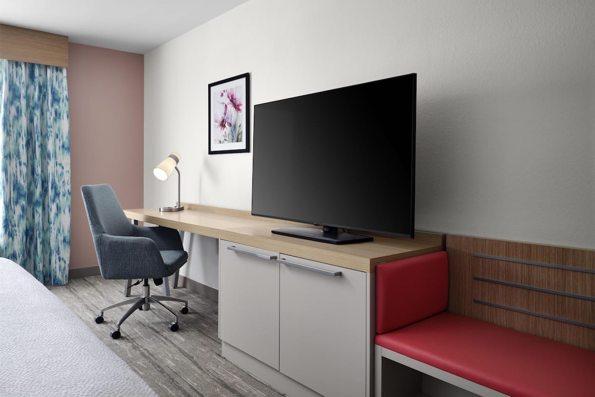 Une chambre d’hôtel avec un lit, un bureau doté d’une télévision, une chaise, une lampe de bureau, un banc rouge et un tableau encadré au mur.