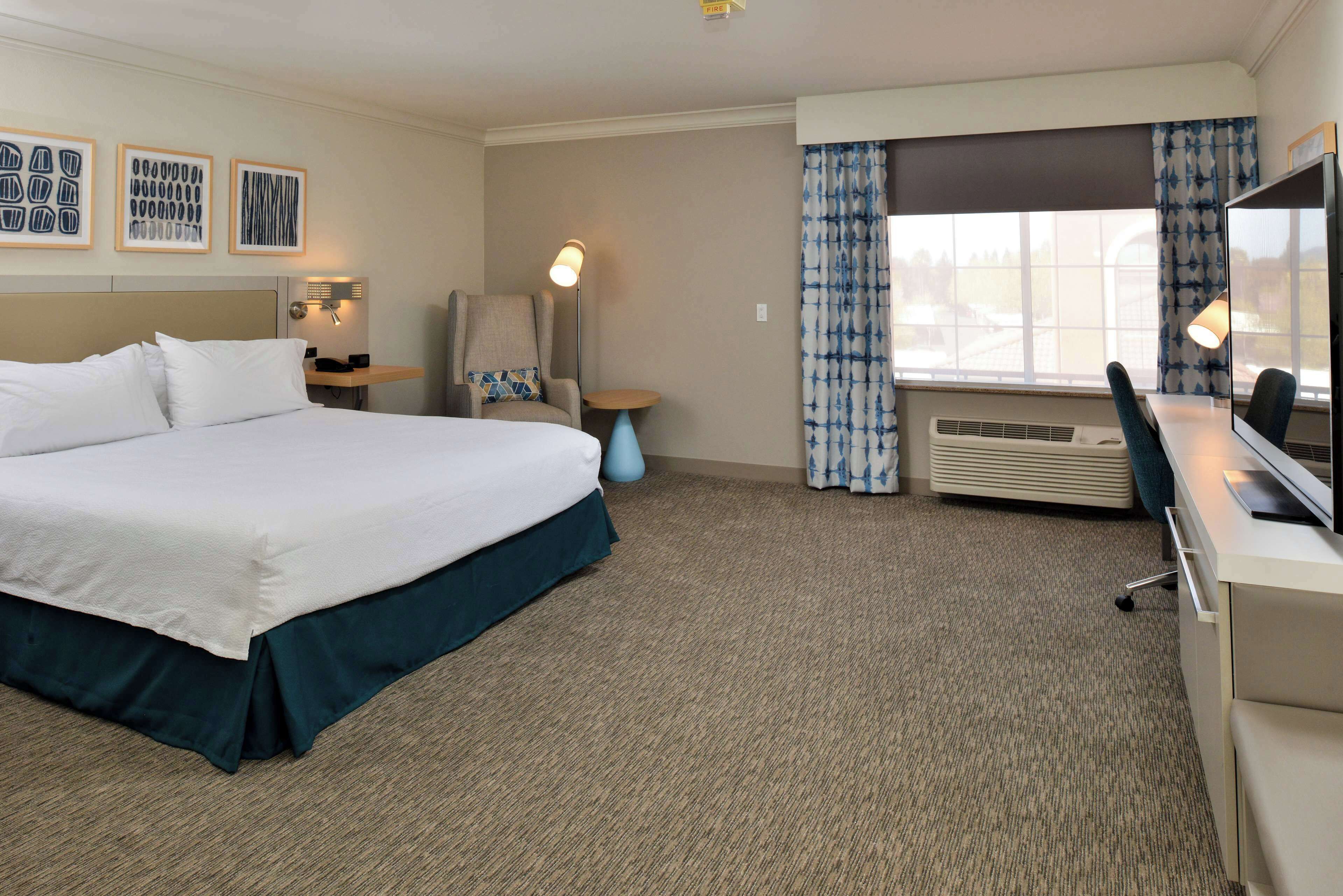 A hotel room with a large bed, a desk with a computer, a chair, a small table, and a window with curtains.