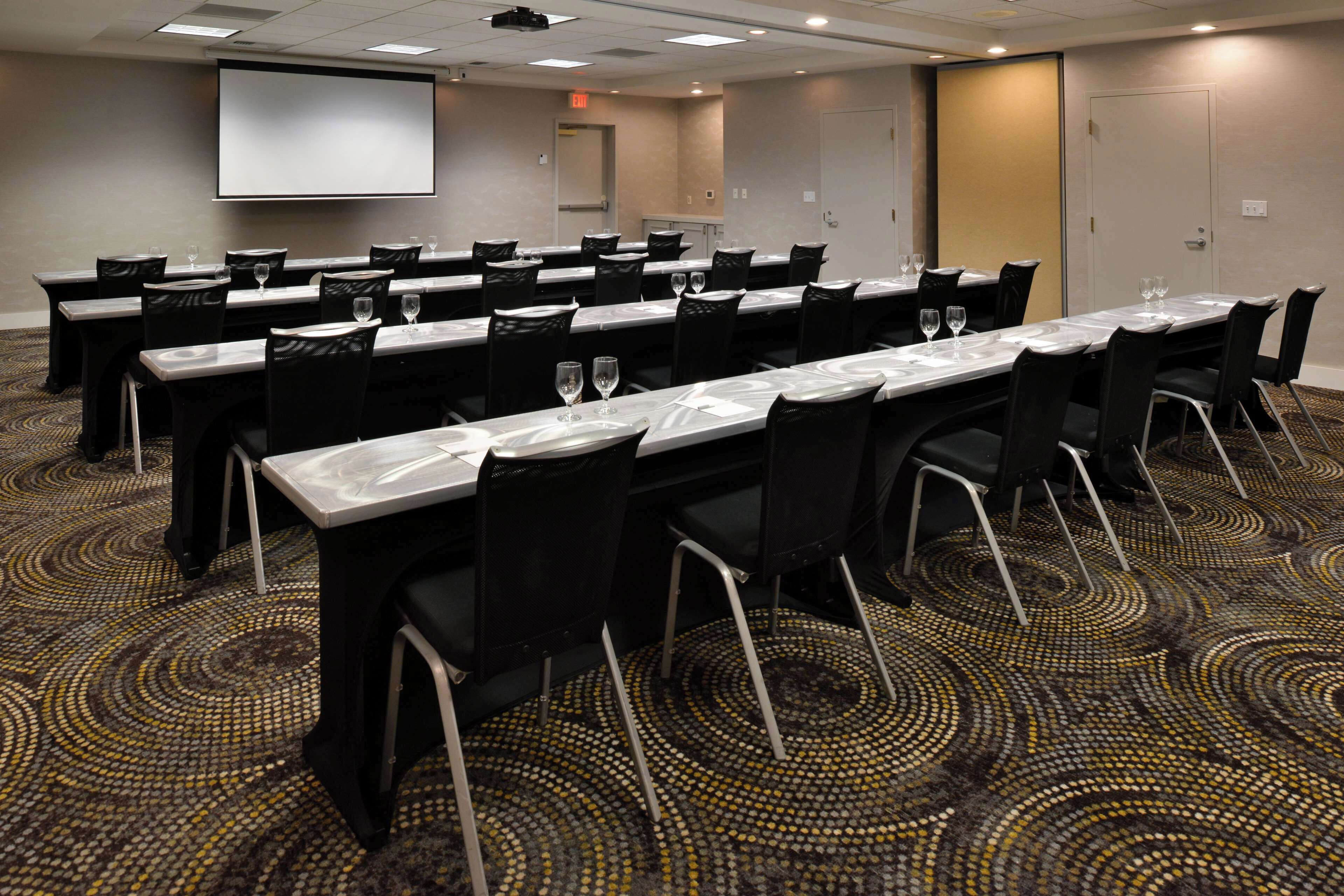 A conference room with multiple tables and chairs, a projector screen, and a carpeted floor.