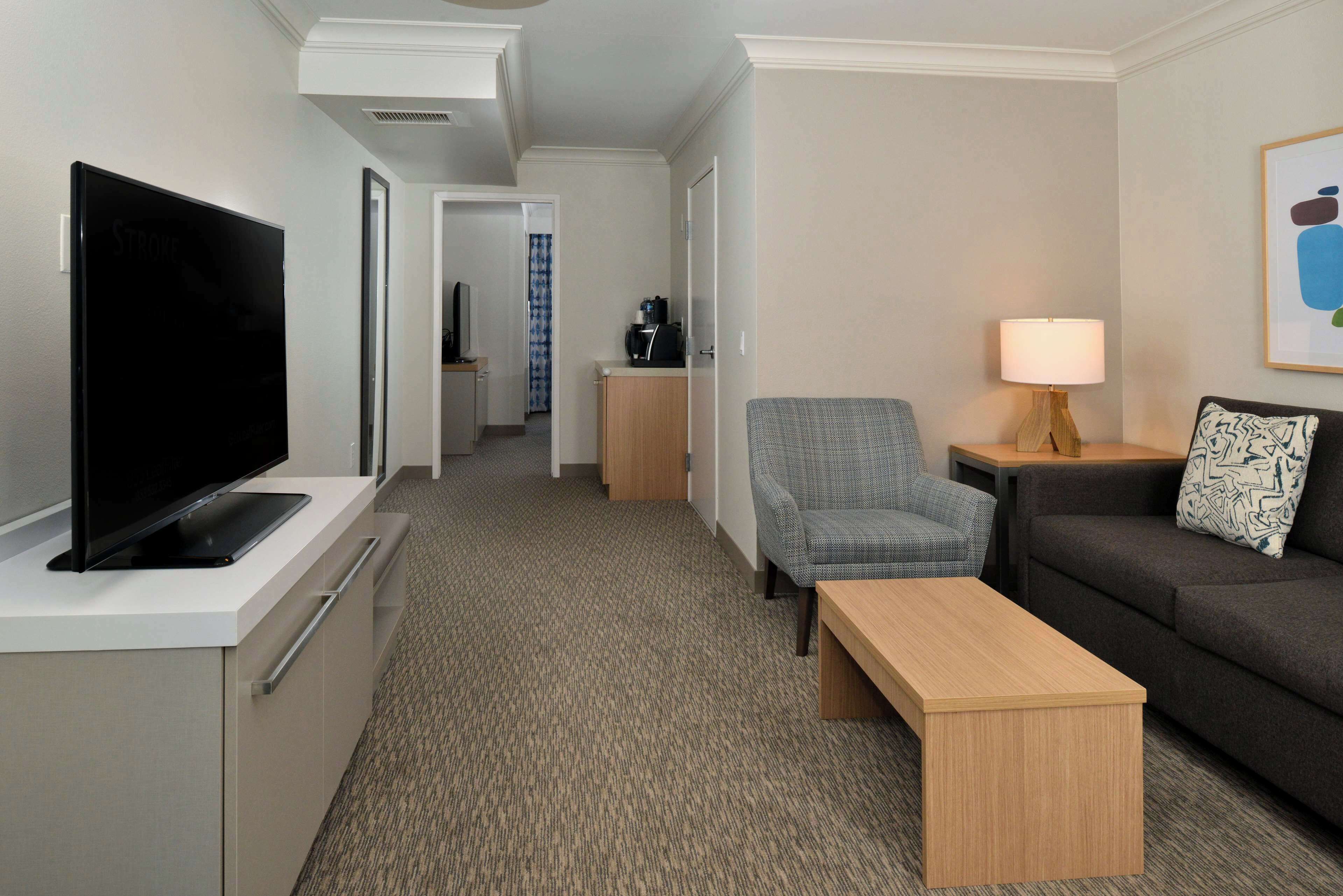 A hotel room with a flat-screen TV on a white stand, a grey armchair, a wooden coffee table, a grey sofa with a patterned pillow, and a lamp on a wooden side table.