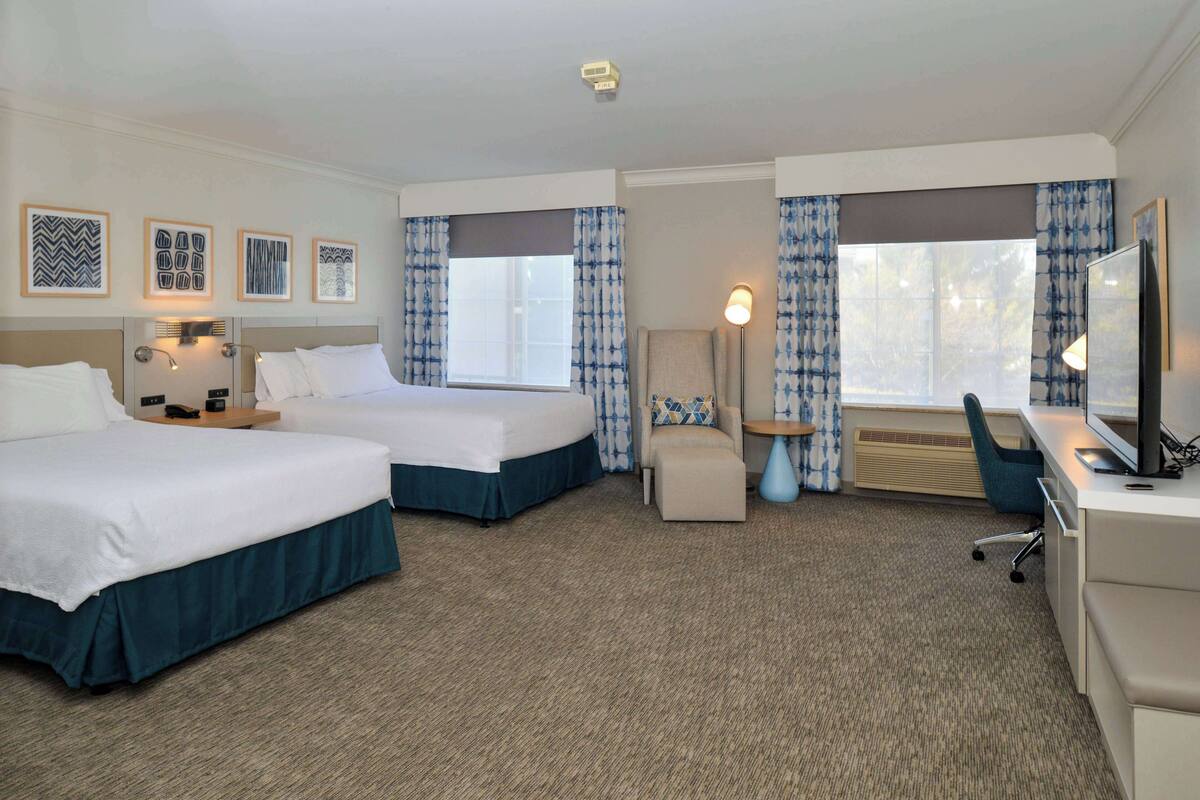 A hotel room with two beds, a desk, a chair, a TV, and a window with curtains.