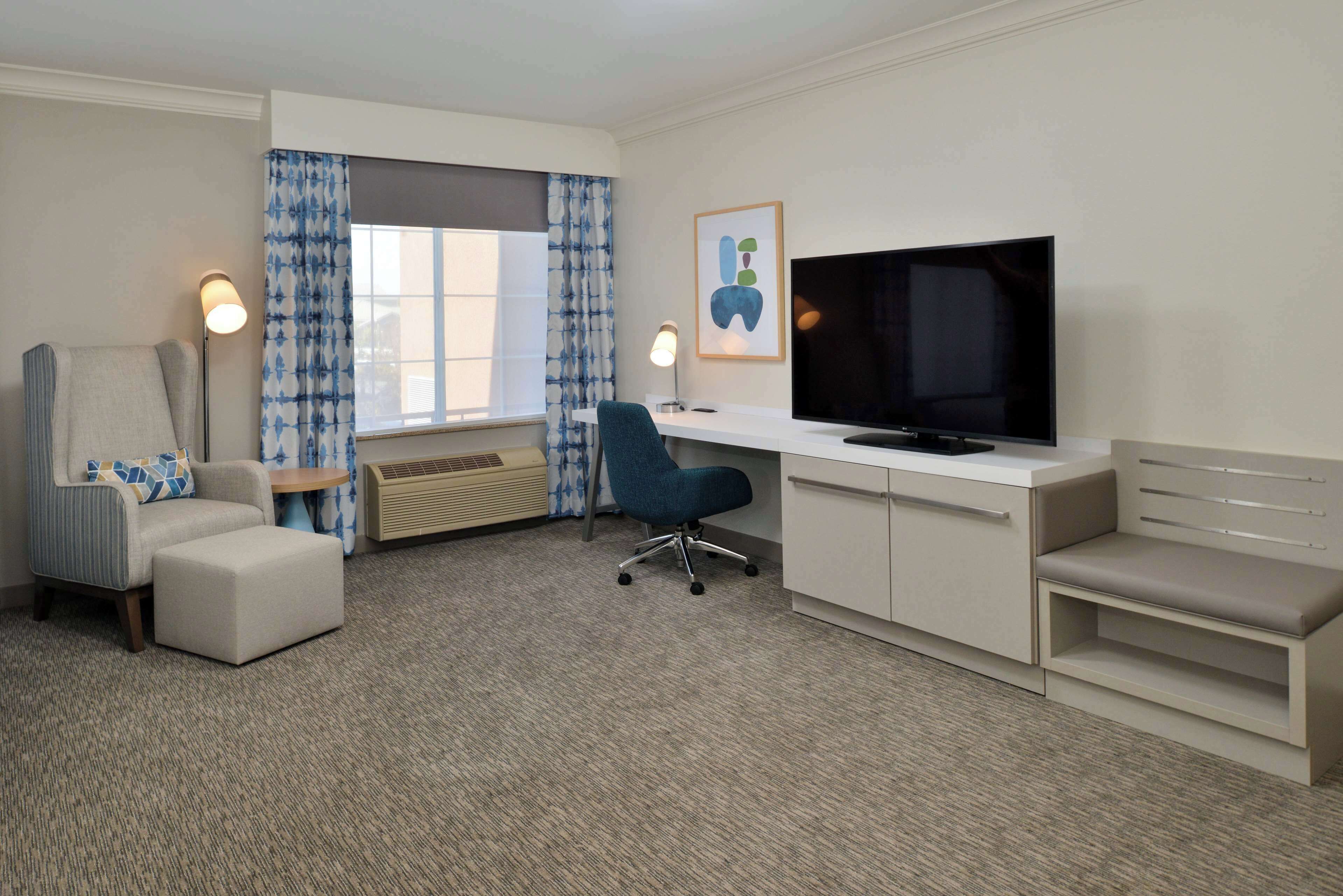 Une chambre d’hôtel dotée d’une grande télévision à écran plat, d’un bureau, d’une chaise, d’un canapé et d’une fenêtre avec des rideaux bleus.