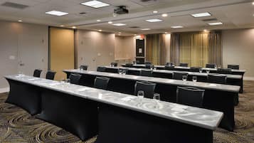 Un salón de conferencias con mesas largas, sillas y una pantalla para proyector.