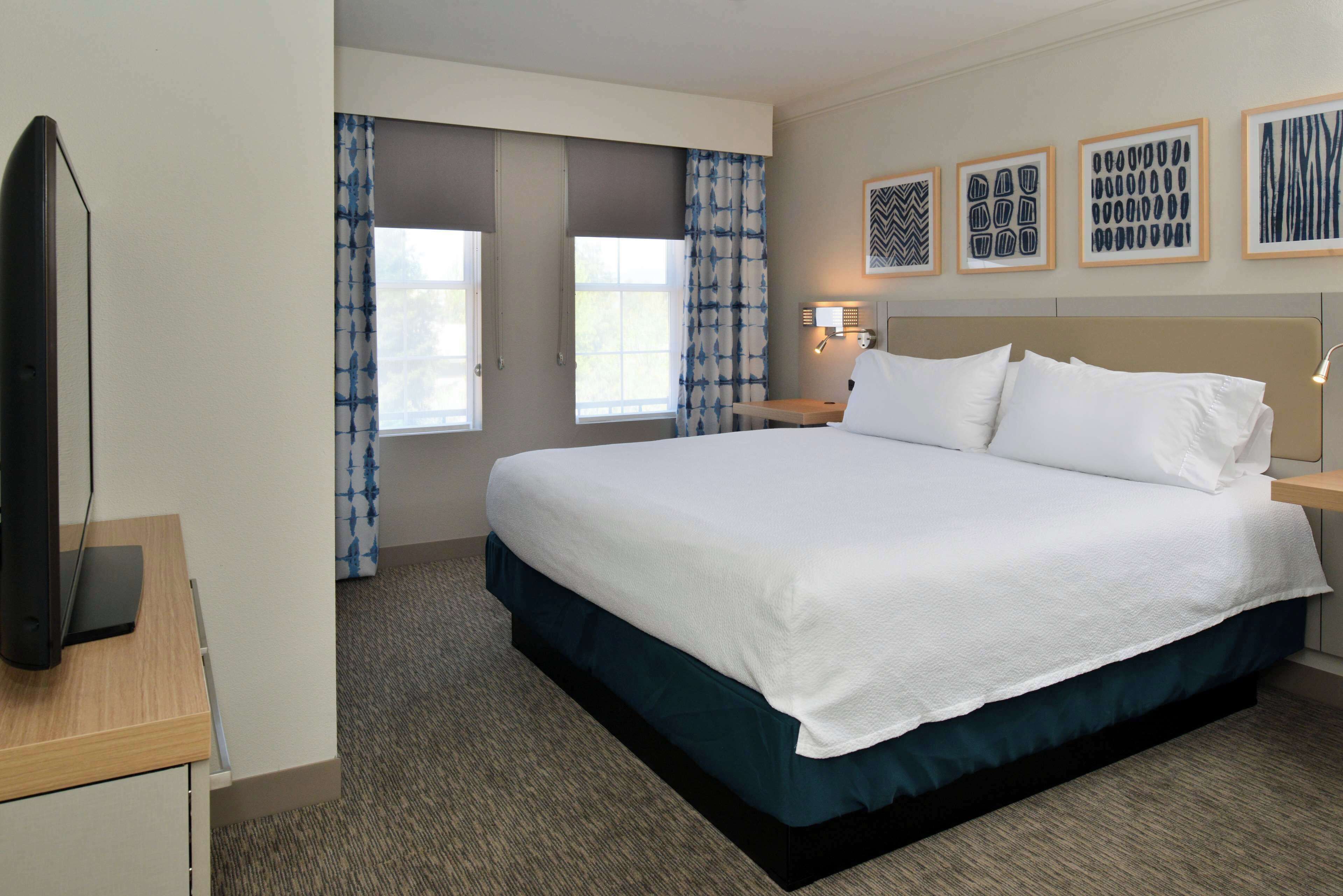 A hotel room with a large bed, a flat-screen TV, a desk with a lamp, and a window with patterned curtains.