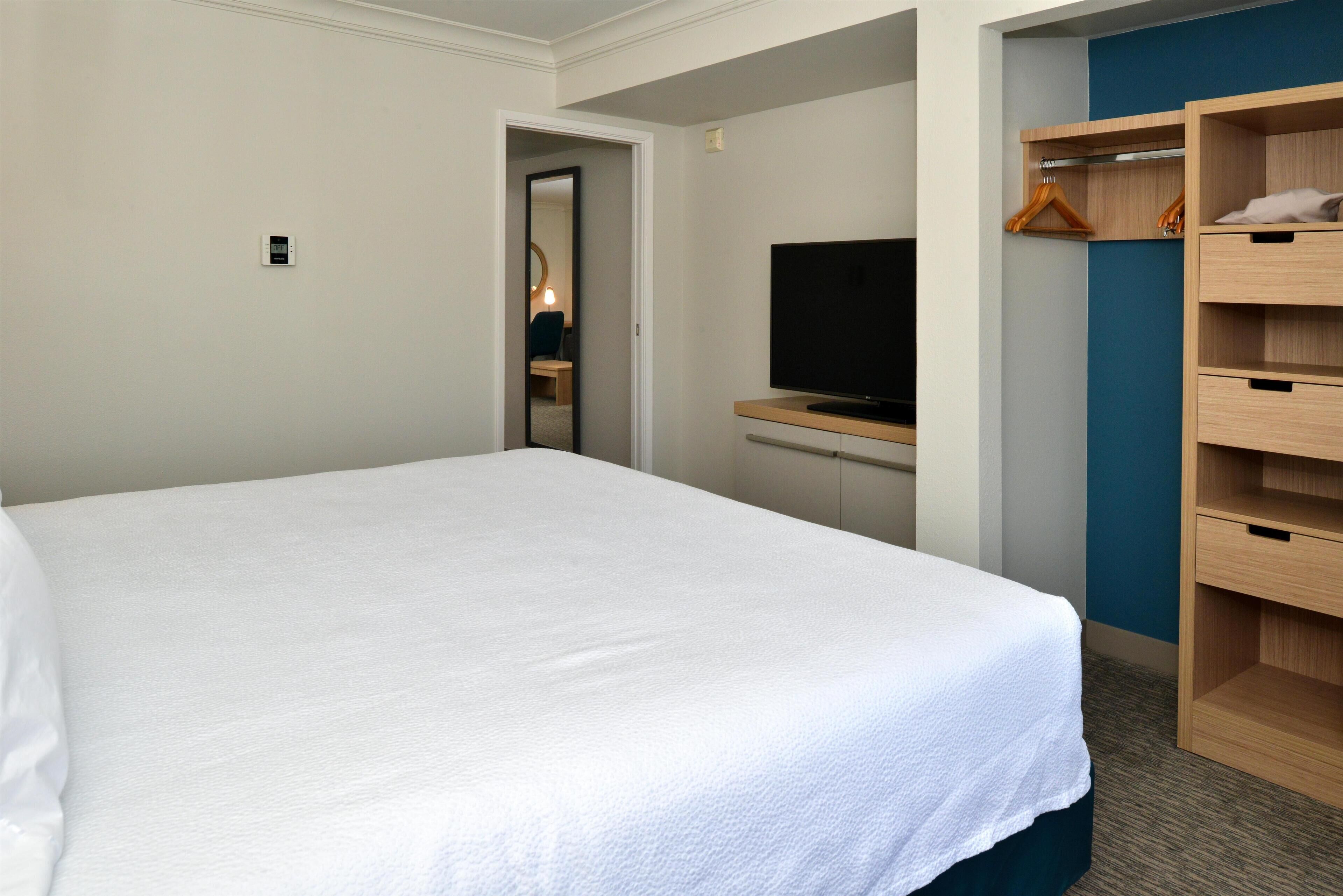 Habitación de hotel con una cama grande, un televisor sobre un mueble, un armario de madera y un espejo en una esquina.