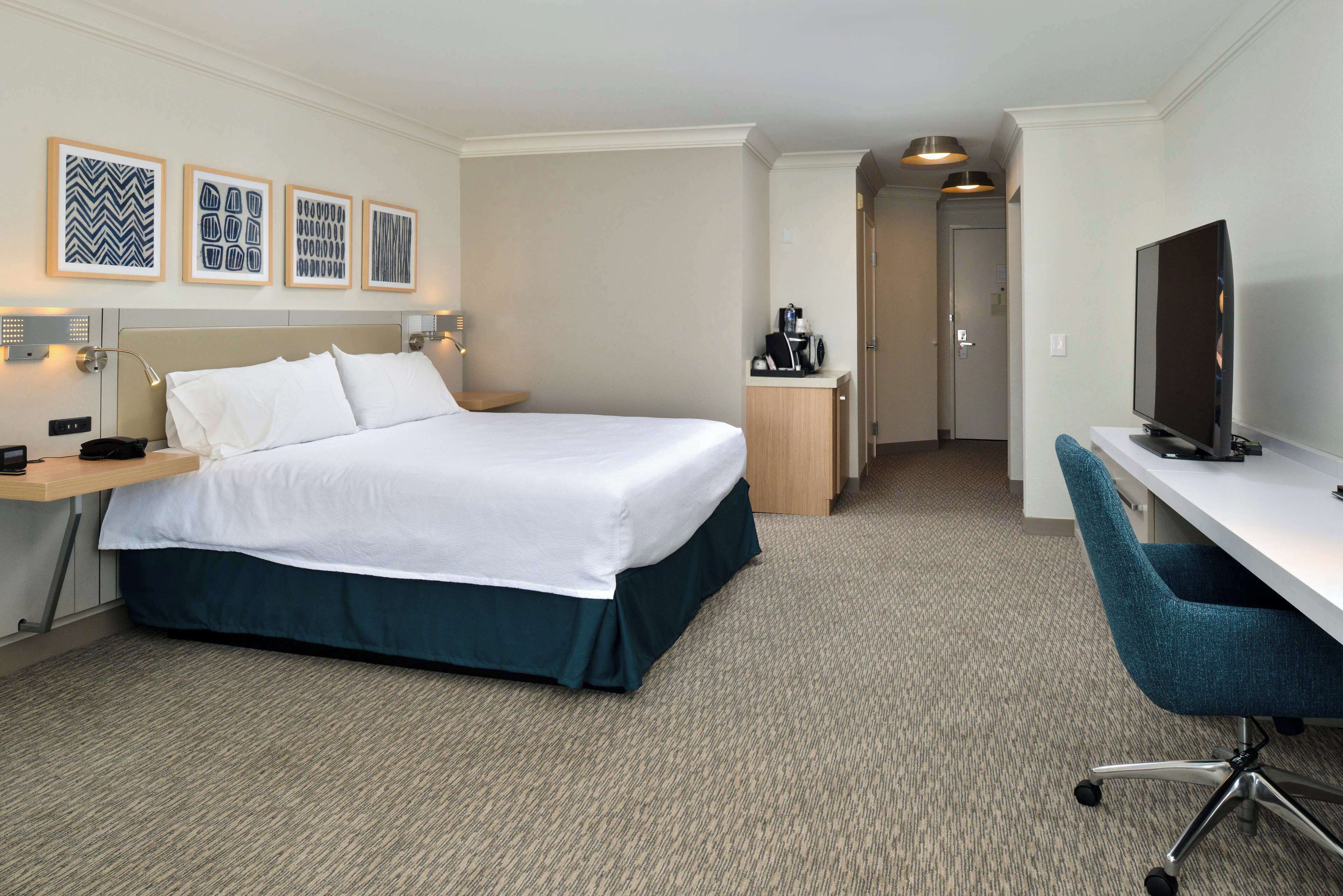 A hotel room with a bed, desk, TV, and framed artwork on the wall.