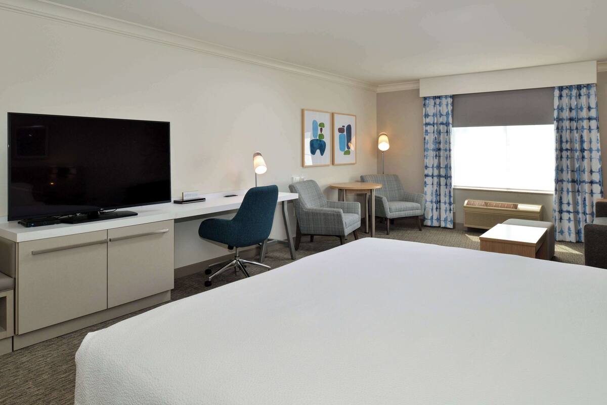 A hotel room with a bed, a desk with a TV, two armchairs, a small table, and a window with curtains.