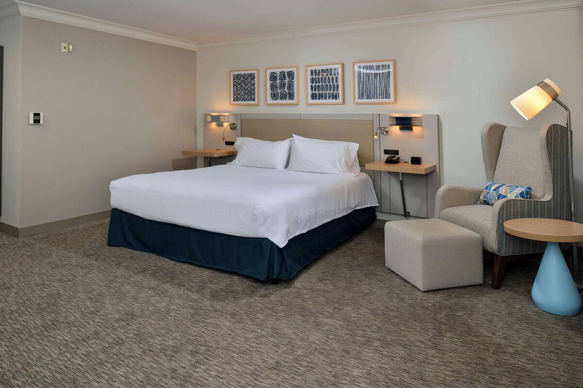 A hotel room with a large bed, bedside tables, a chair, a small table, and wall art.