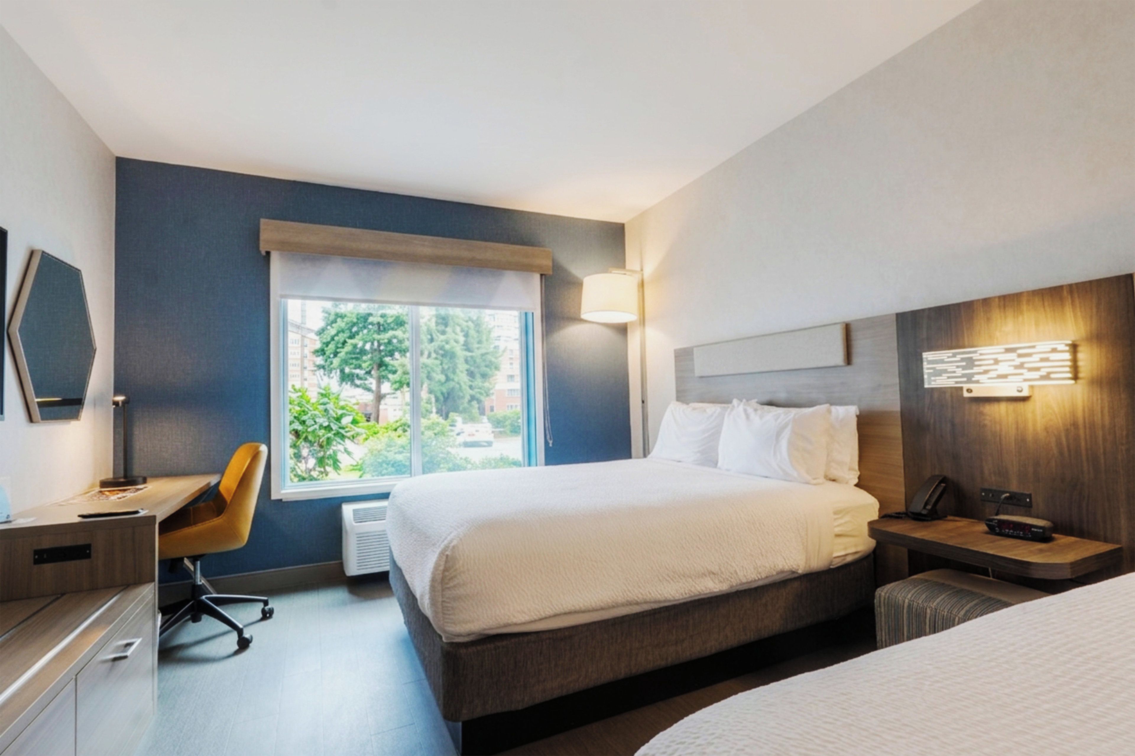 Ein Hotelzimmer mit Bett, Nachttisch, Lampe, Stuhl, Schreibtisch und einem Fenster mit Blick auf Grünflächen.