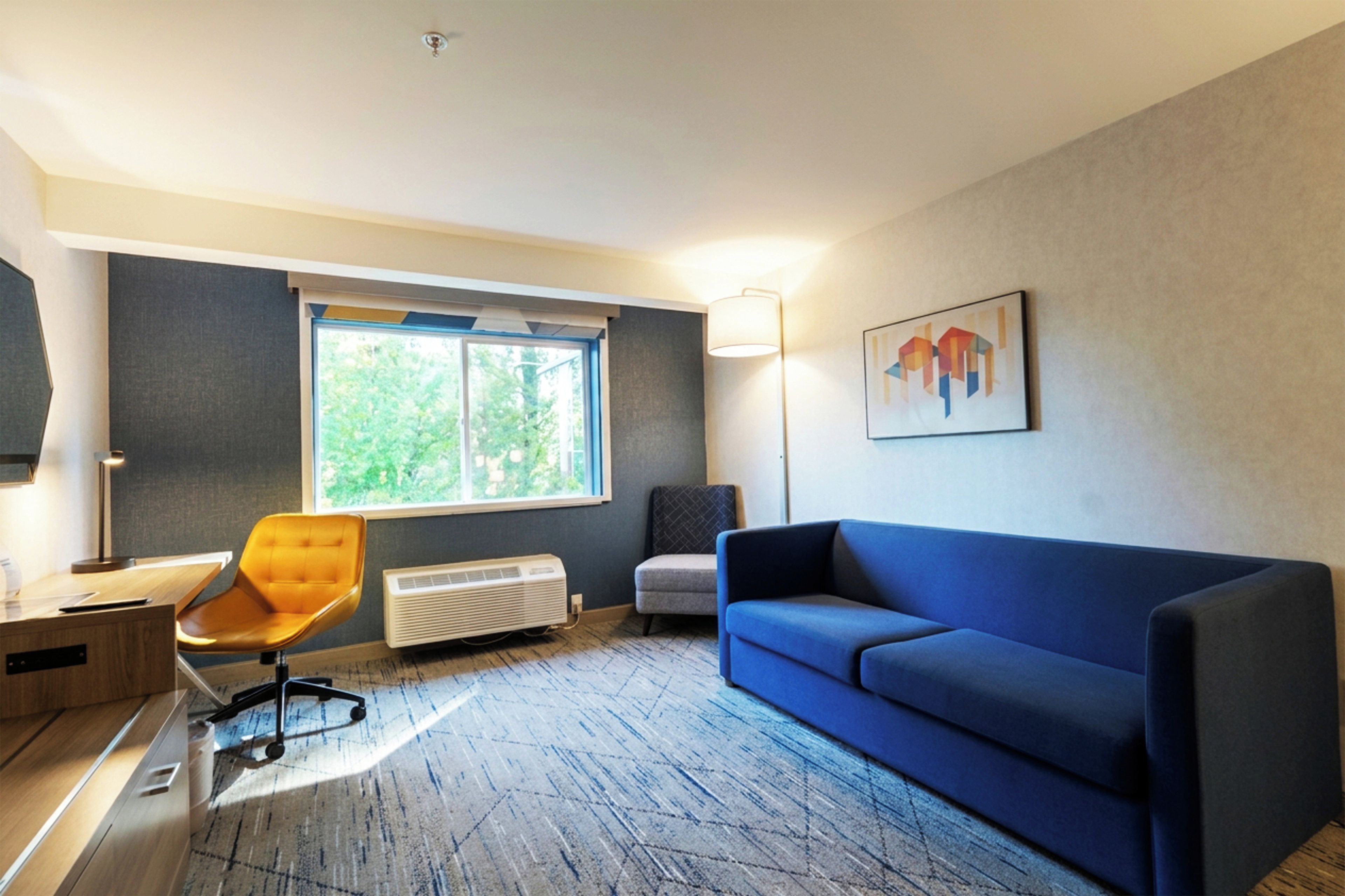 A modern living room with a blue sofa, a yellow armchair, a window with a view of greenery, and a painting on the wall.
