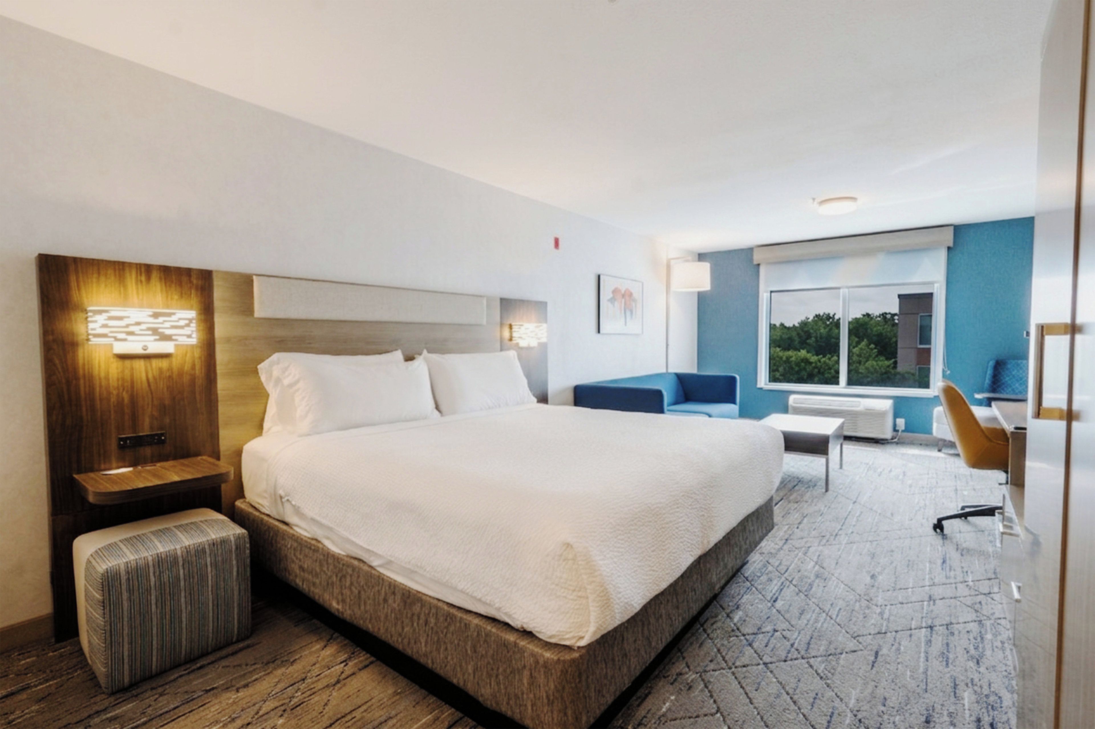 Habitación de hotel con una cama grande, cabecera de madera, mesita de noche, un banco, un escritorio, una silla y una ventana con vistas.