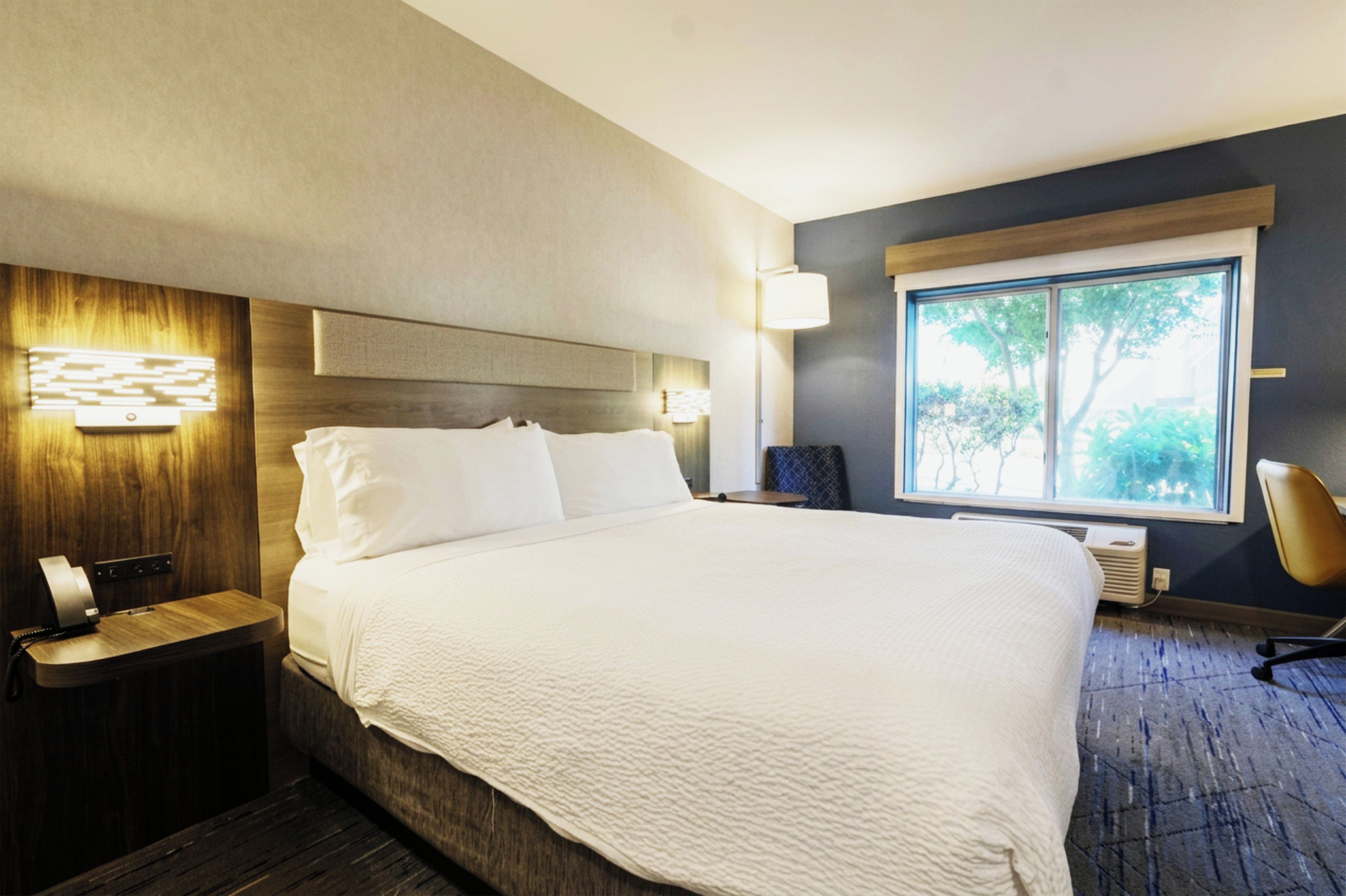 A hotel room with a large bed, bedside tables, a desk, a chair, a window with a view, and a wall-mounted lamp.