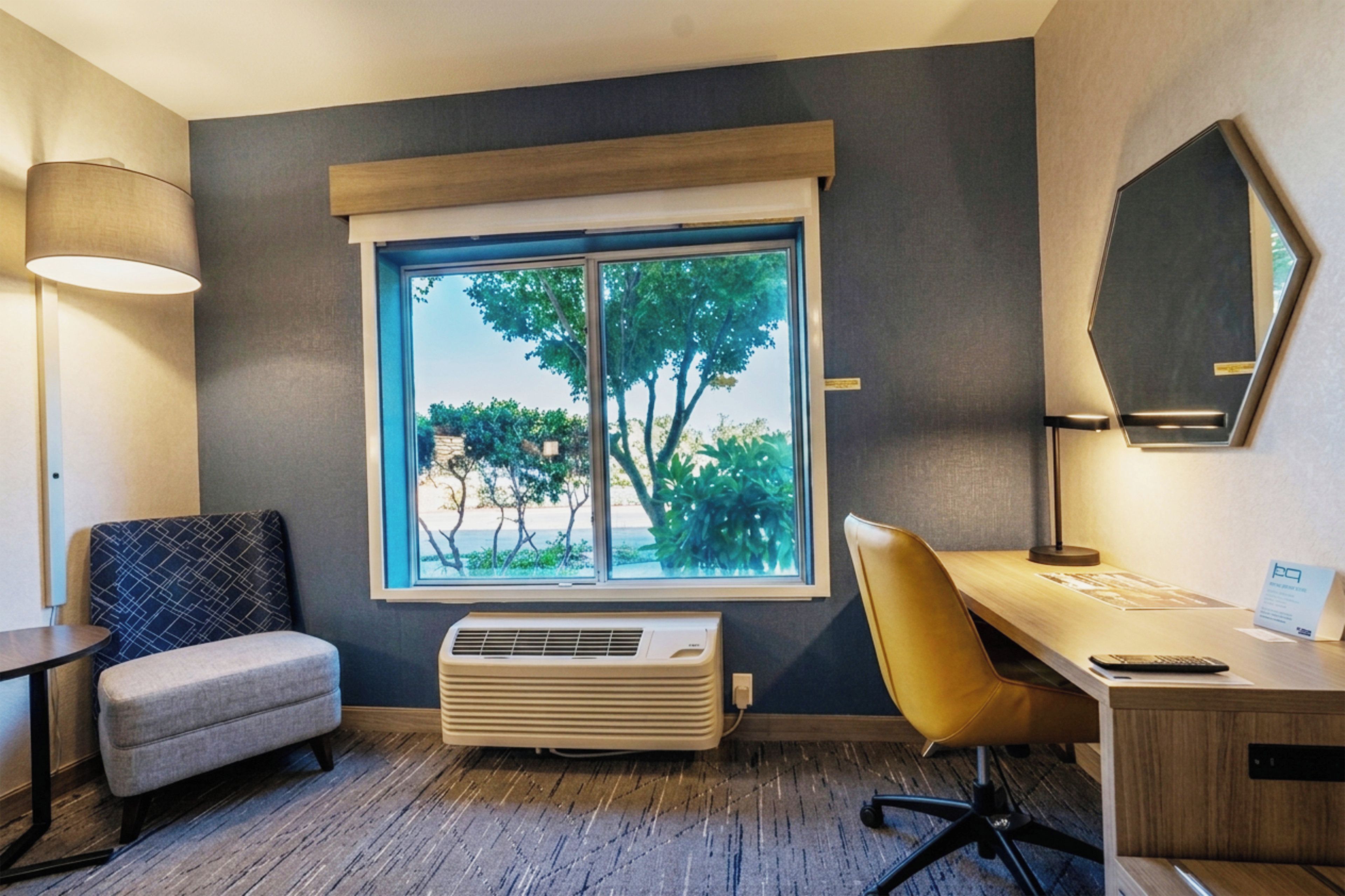 Una habitación con escritorio, silla y un ventanal con vista a árboles.