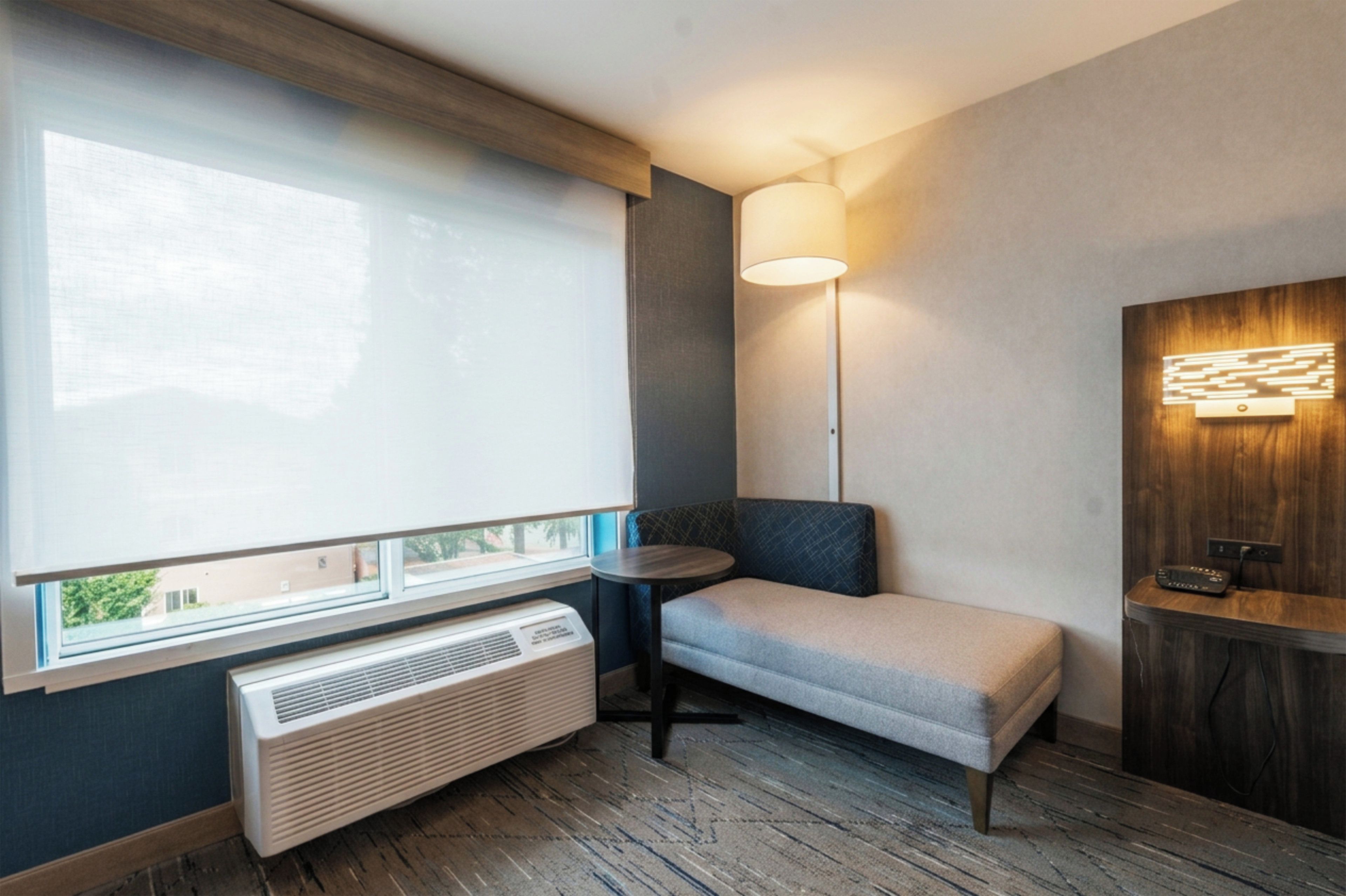 A modern room with a large window, a standing heater, a small table, a bench, and a wall-mounted lamp.