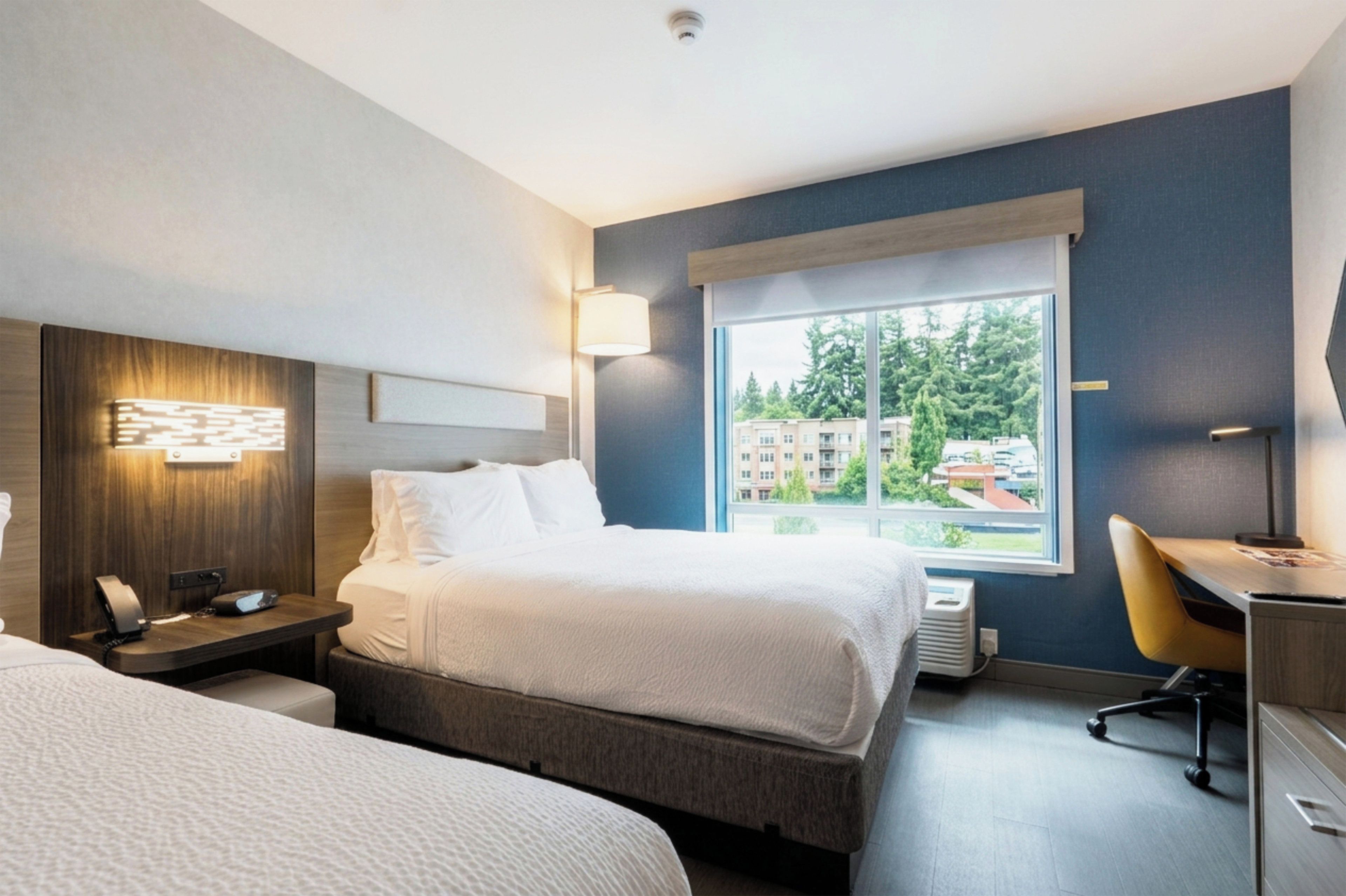 Ein Hotelzimmer mit Bett, Nachttisch, Lampe und Blick auf Gebäude durch das Fenster.