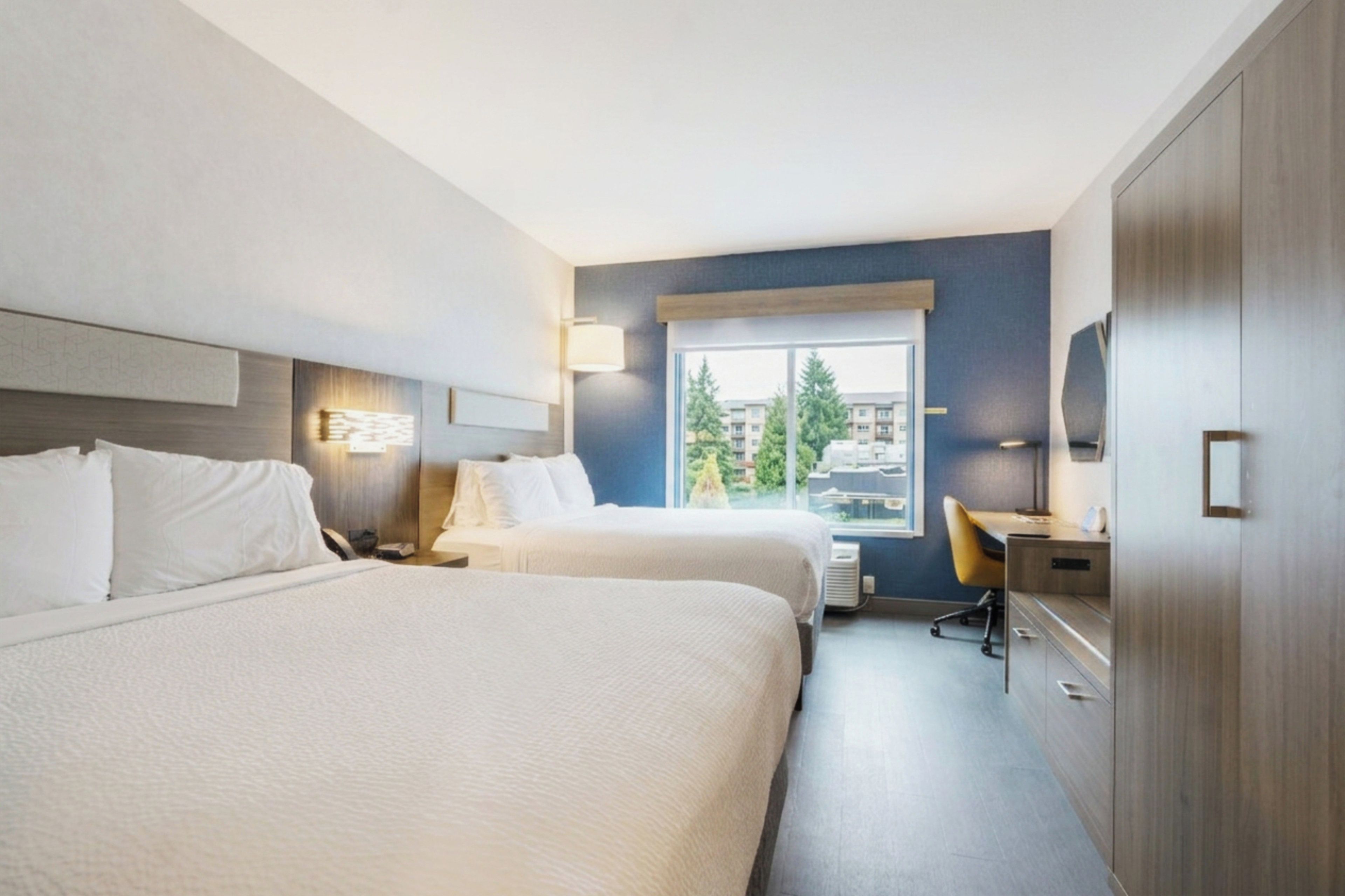 A hotel room with two beds, a desk, a chair, a wardrobe, and a window with a view of buildings and trees.